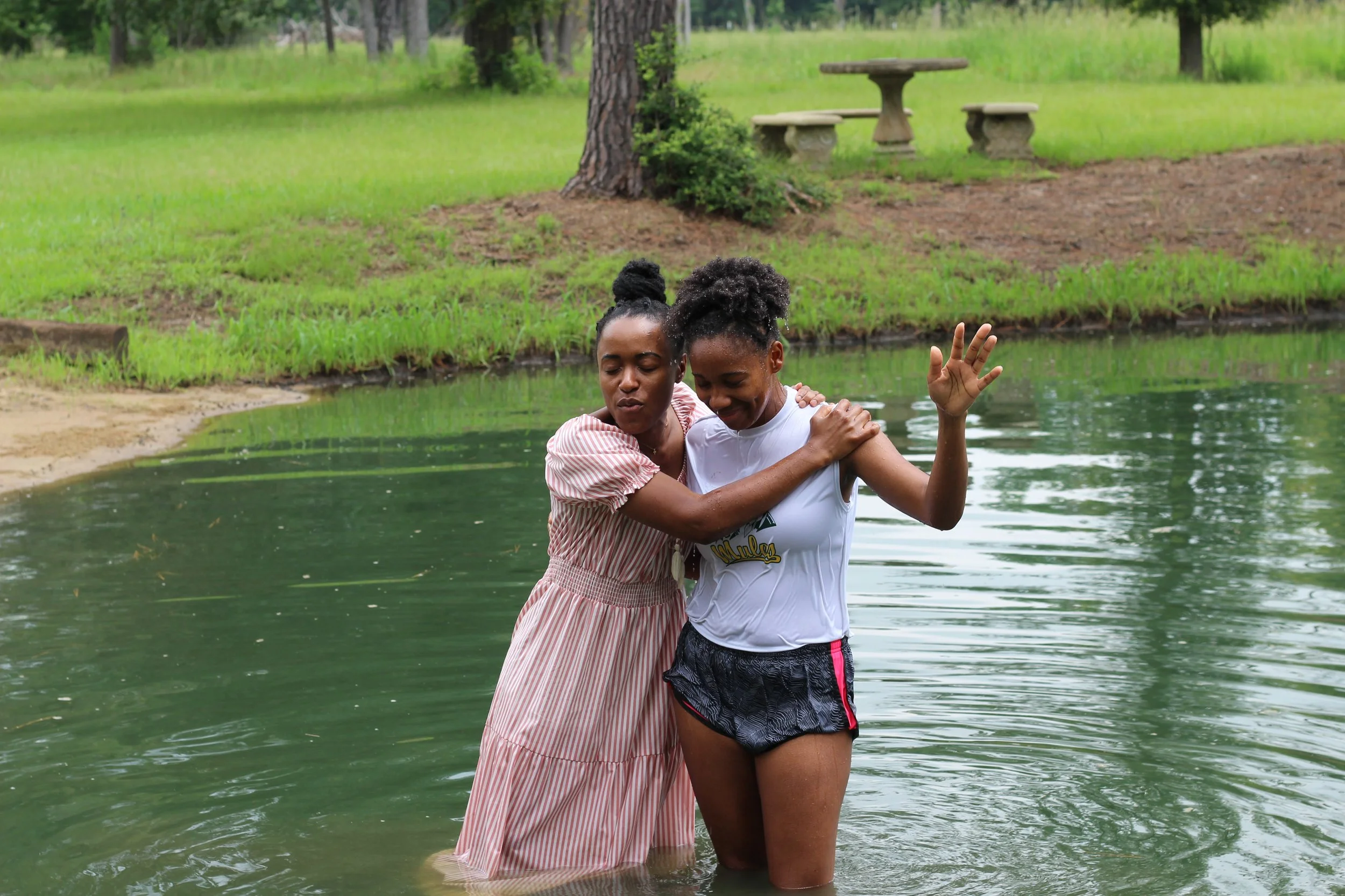 Two women standing in a pond, embracing each other with happy expressions, surrounded by green grass and trees in the background.