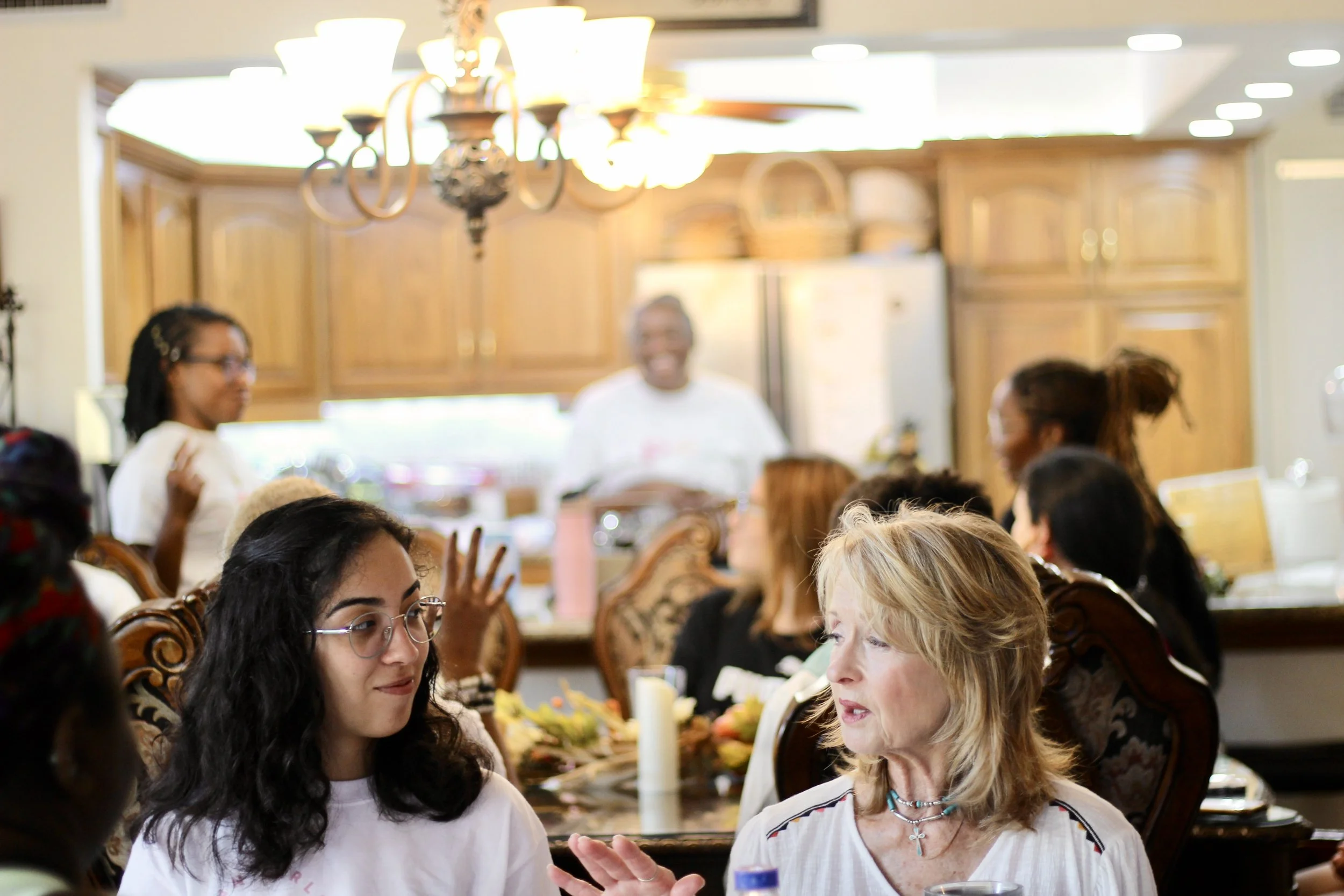 Group of women gathered around a dinner table engaged in conversation, with a woman in the background smiling in a kitchen setting.