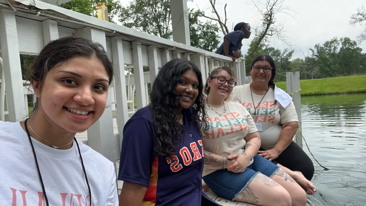 Five women sitting by a pond, smiling for a photo, with a child leaning on a railing behind them.