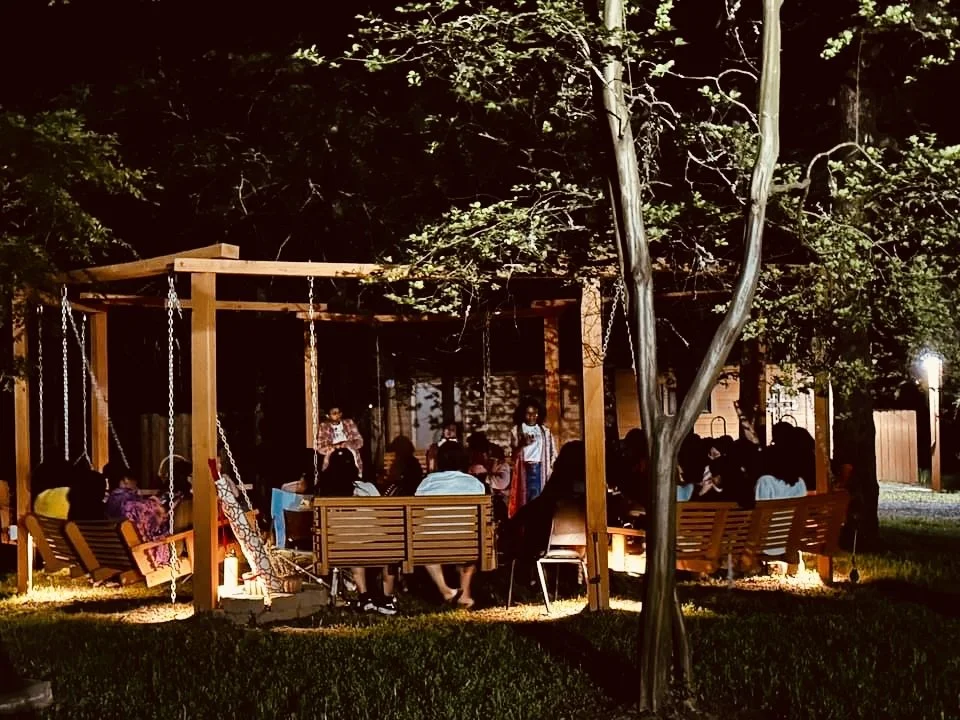 A group of people gathered around a wooden pavilion with swings, in a park at night, illuminated by outdoor lights.