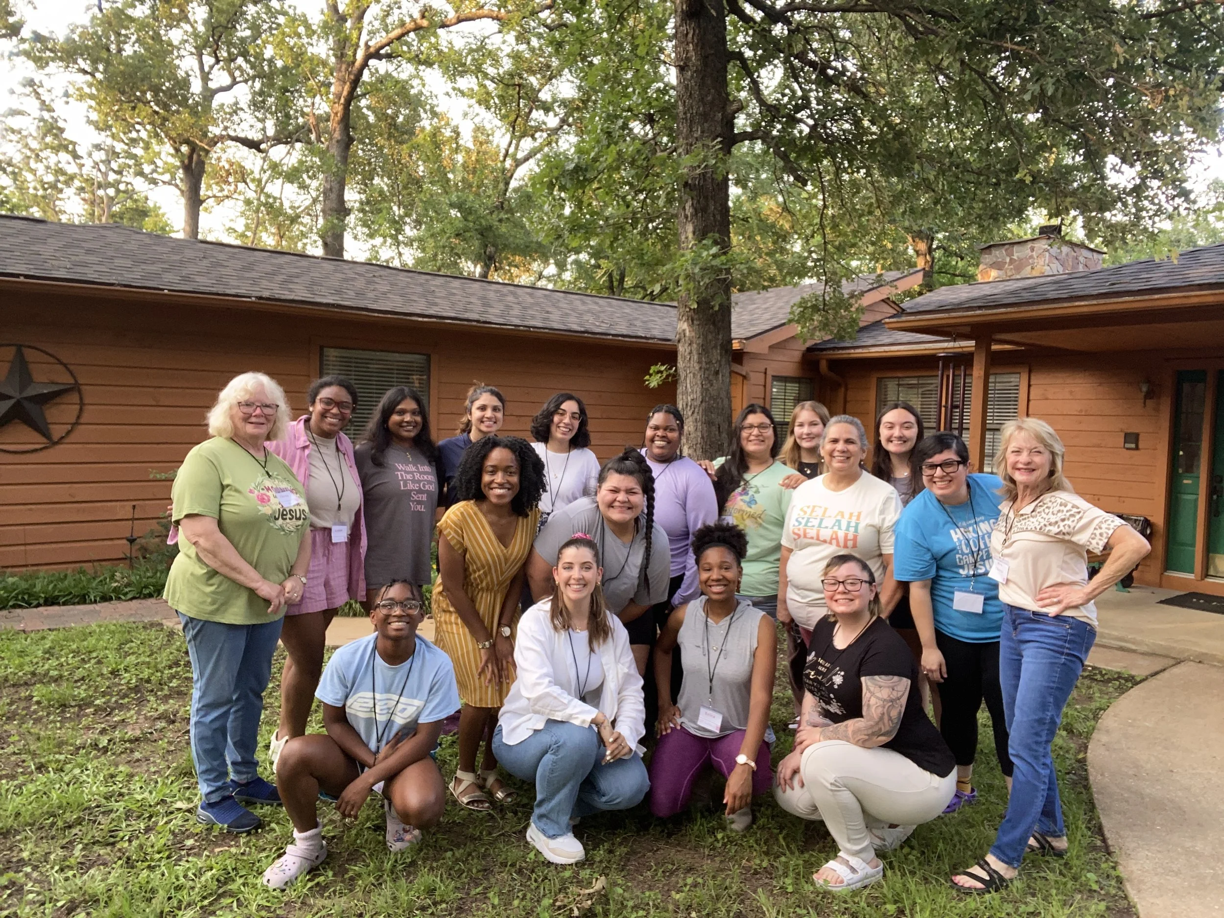 Group of diverse women standing outside in front of a wooden building with a large tree overhead, posing for a photo.
