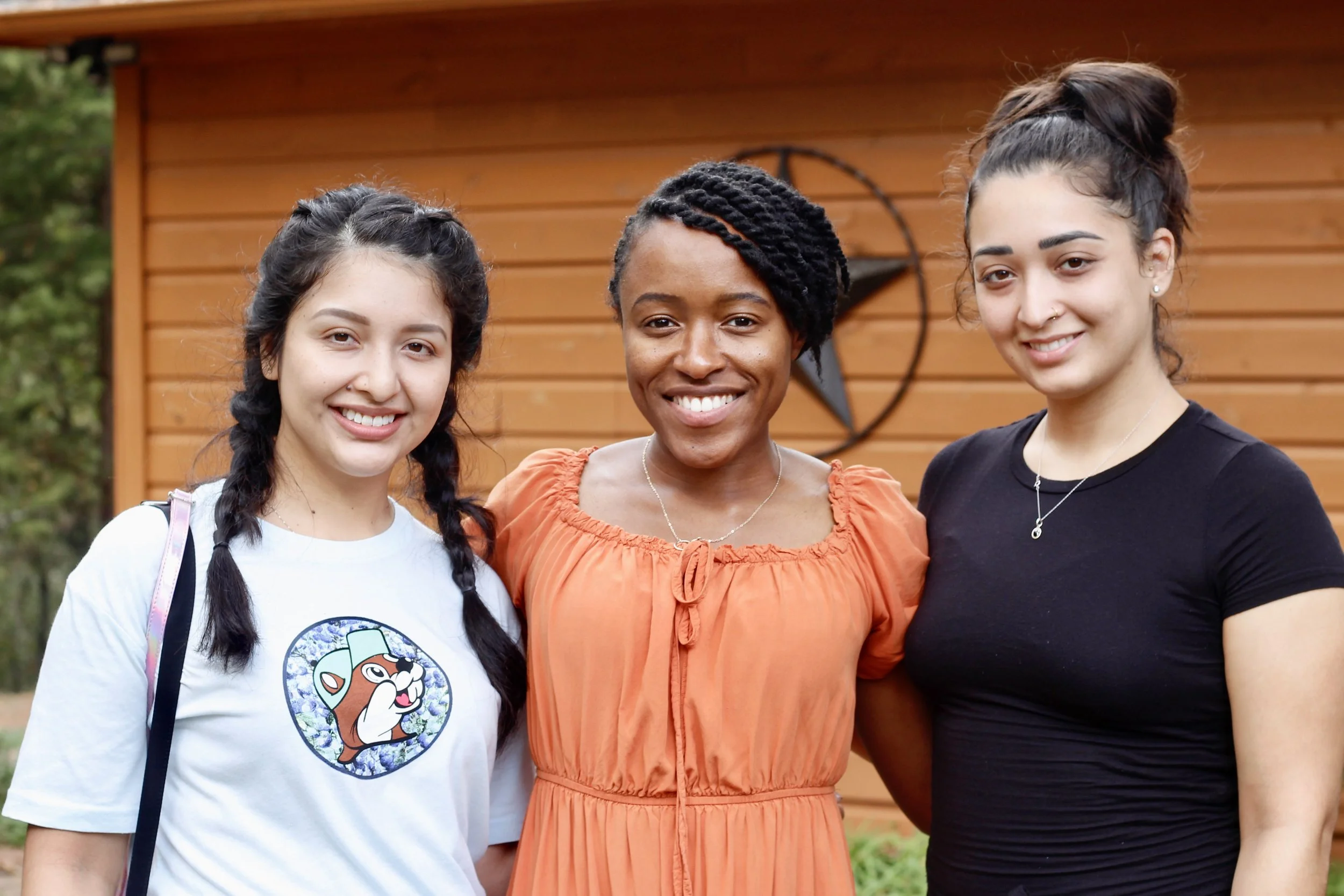 Three women standing outdoors and smiling, with wooden wall and star decoration in the background.