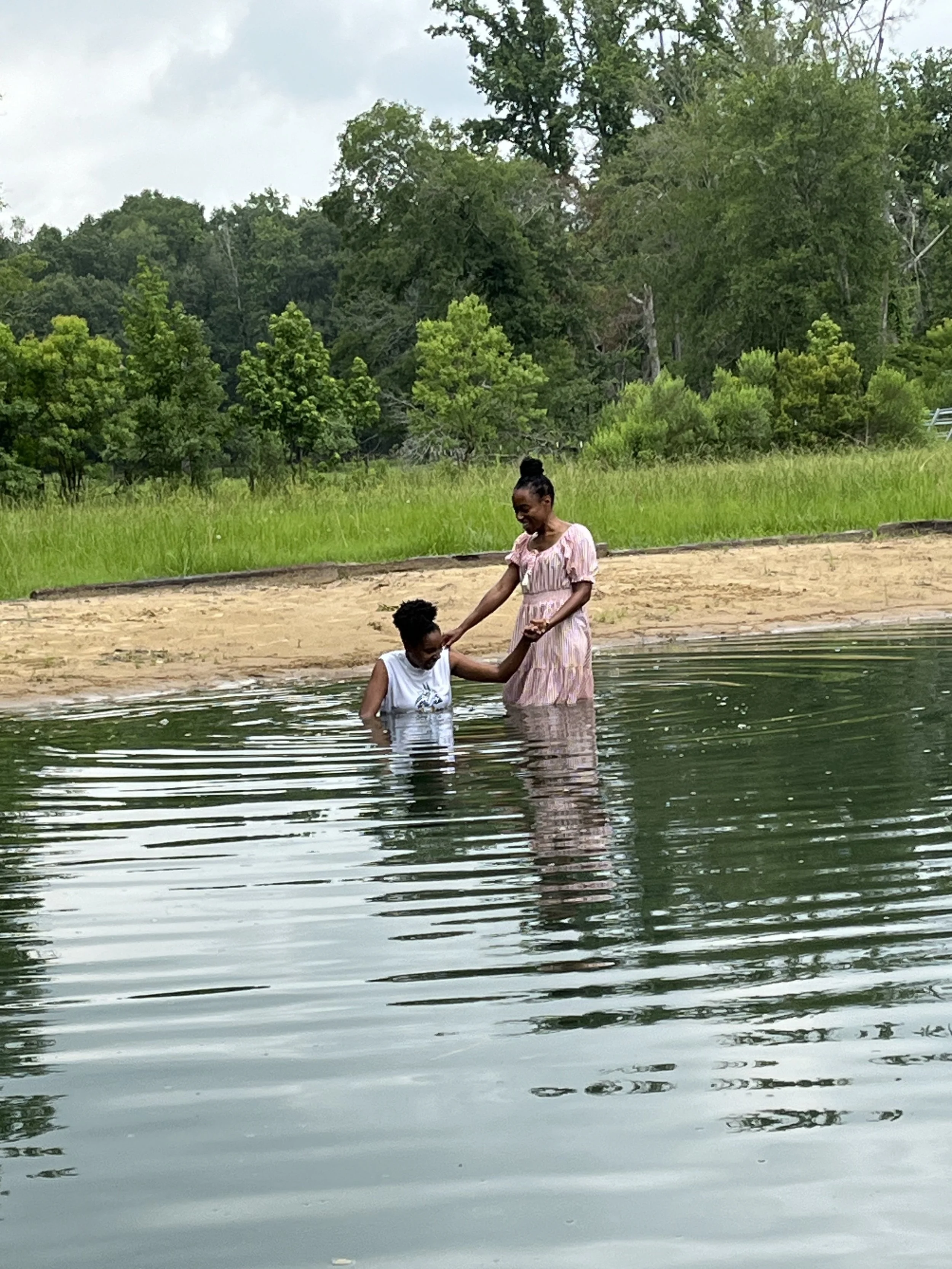 A woman and a girl standing in a body of water, holding hands and smiling, with trees and grass in the background.
