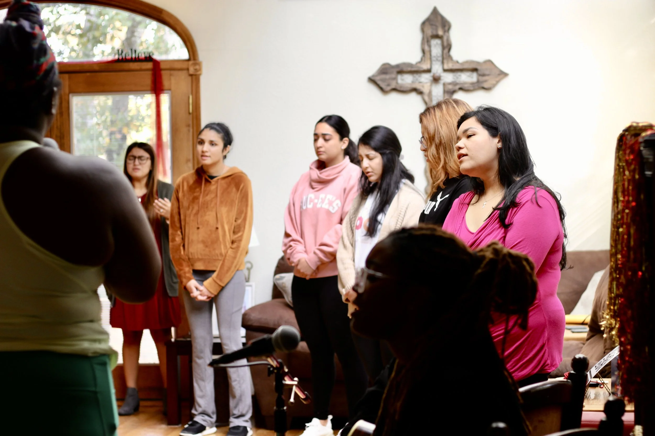 A group of women standing together indoors, with some looking down and others looking ahead, in what appears to be a prayer or meditation session.