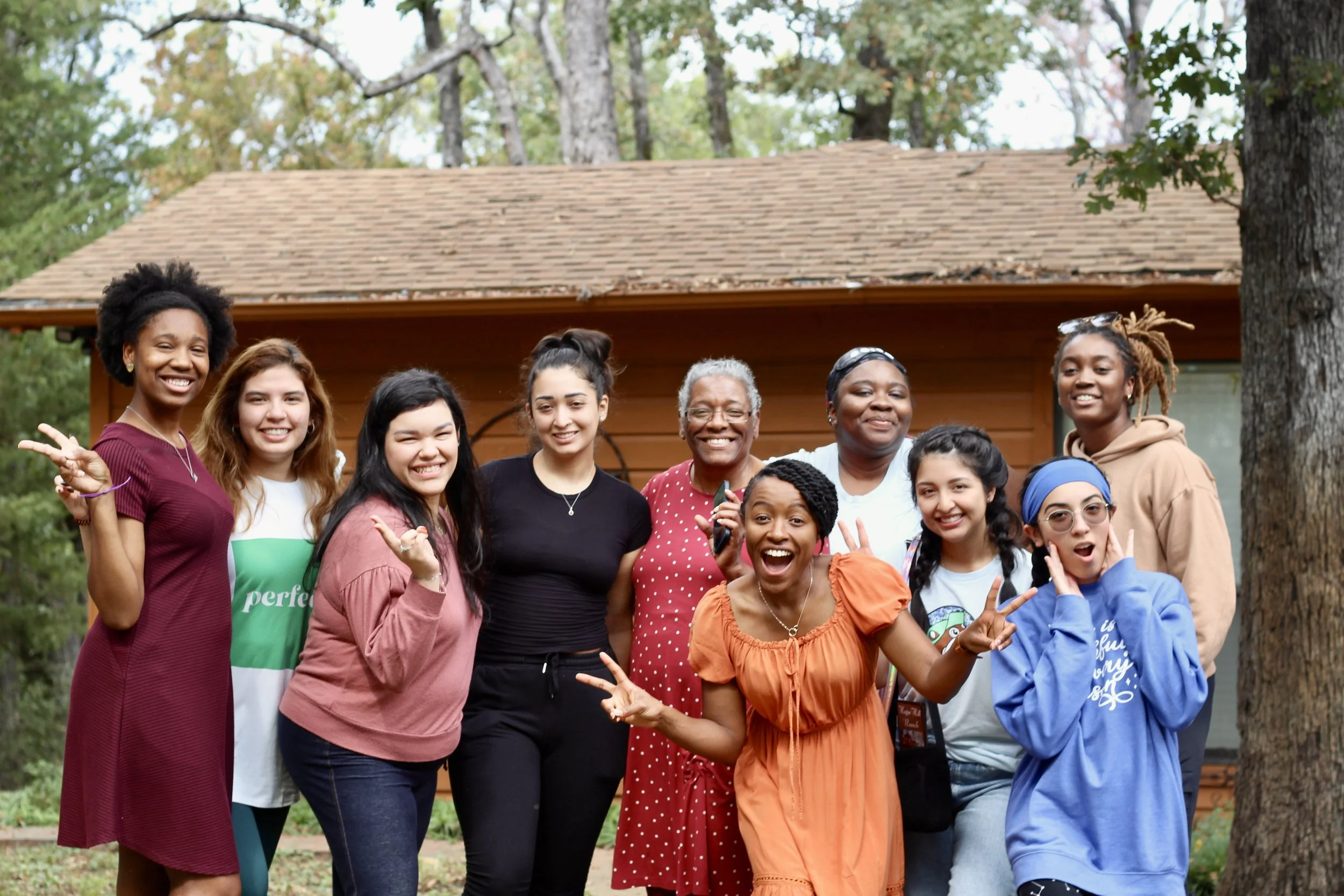 Group of diverse women and girls smiling and posing outdoors in front of a wooden house.