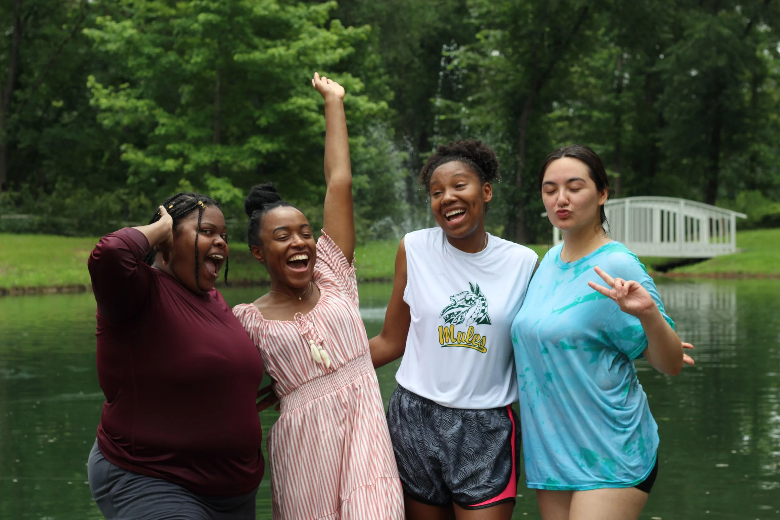 Four women standing outdoors by a pond, smiling and celebrating, with trees and a small white bridge in the background.