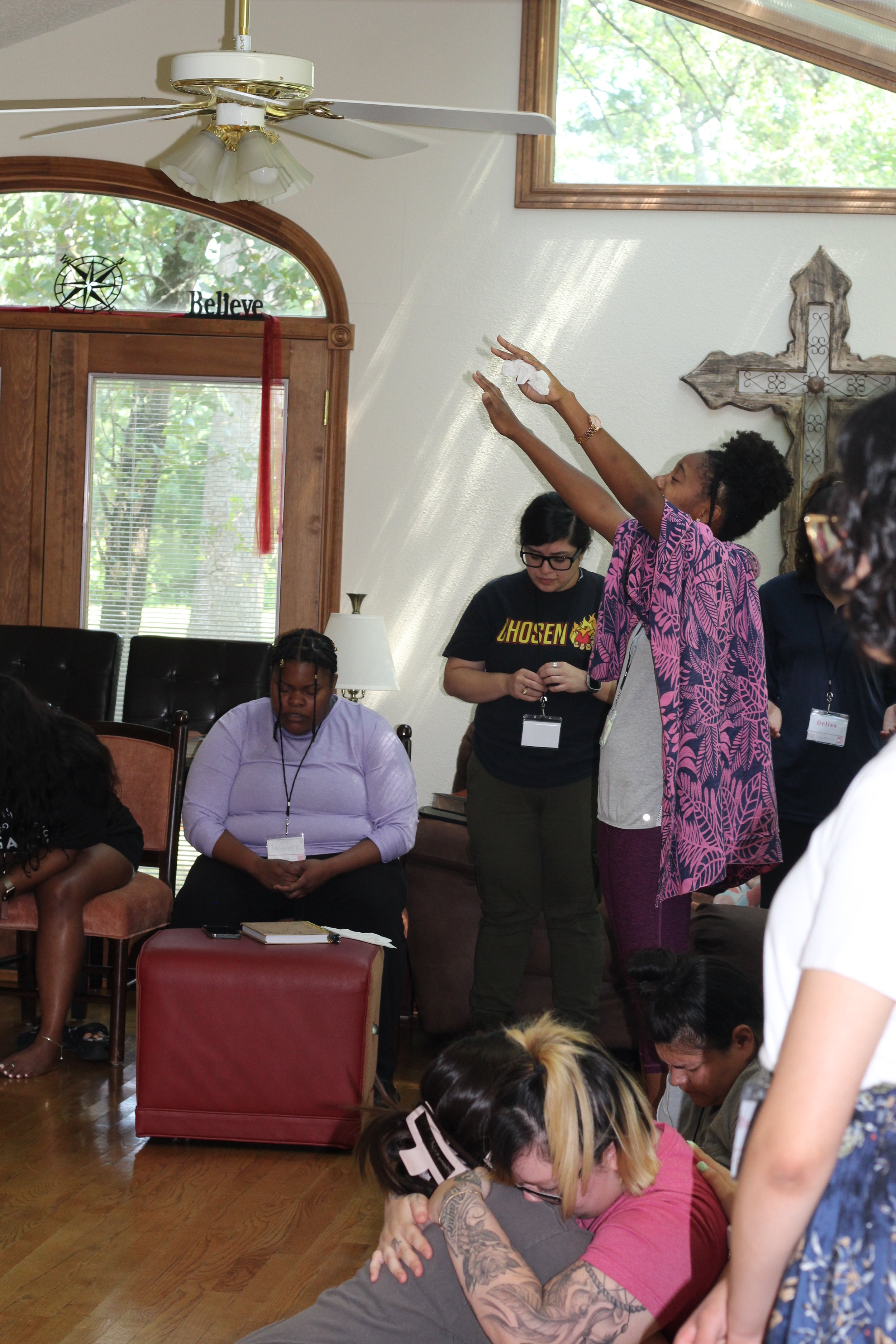 A group of women participating in a prayer circle inside a room with wooden floors, a cross on the wall, and a ceiling fan. Some women are standing, some kneeling or sitting, with one woman in pink reaching upward, and others with eyes closed or bowe