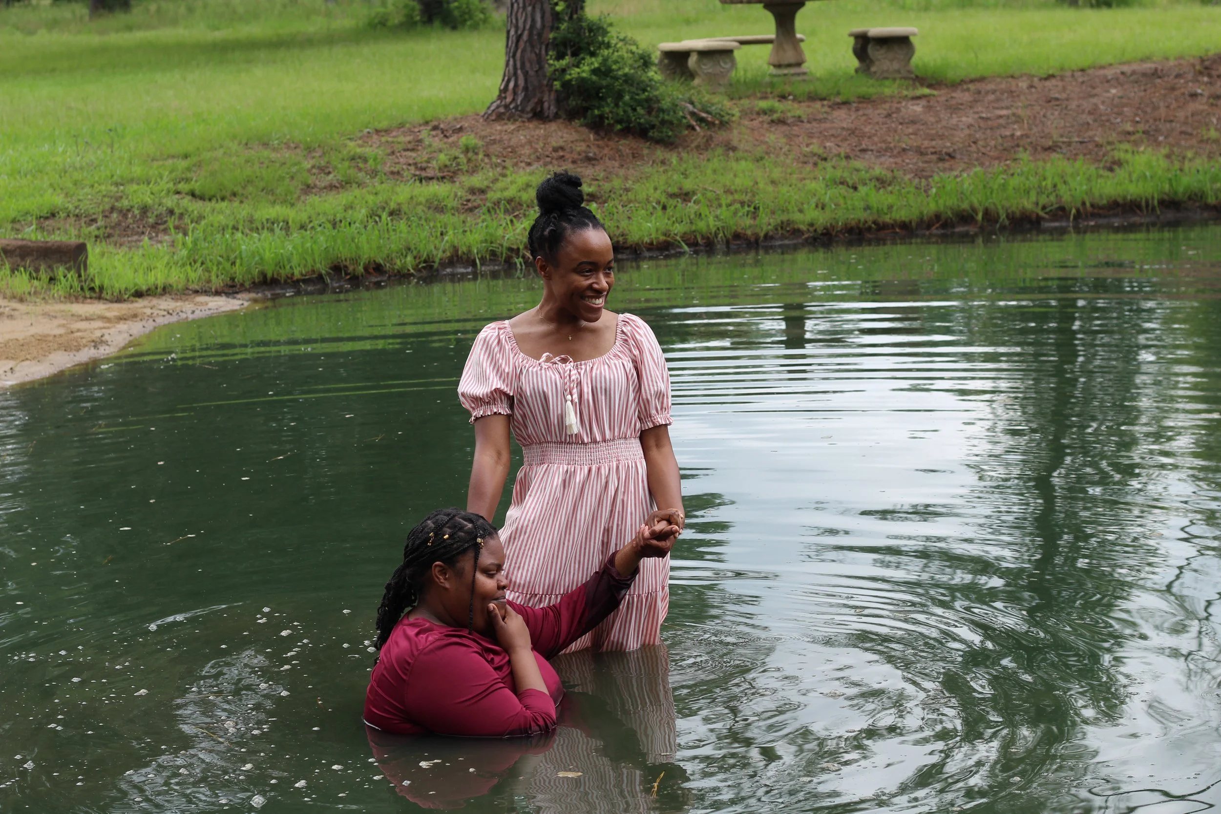 Two women are standing in a peaceful pond surrounded by green grass and trees. One woman is standing and smiling, wearing a pink and white striped dress, while the other woman is kneeling, resting her head on her hand, and wearing a dark red shirt.