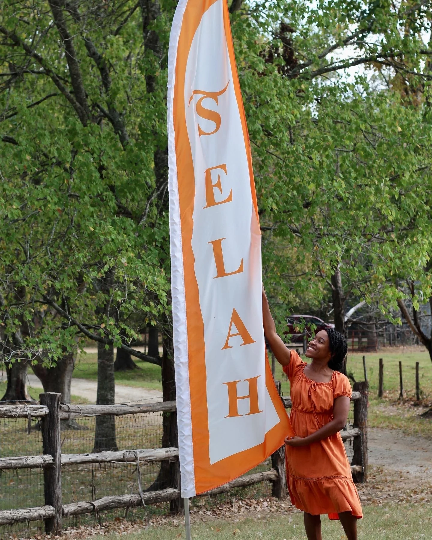 A woman in an orange dress smiling while holding a large flag with the word 'SELAH' written vertically in orange letters outside near trees and a wooden fence.