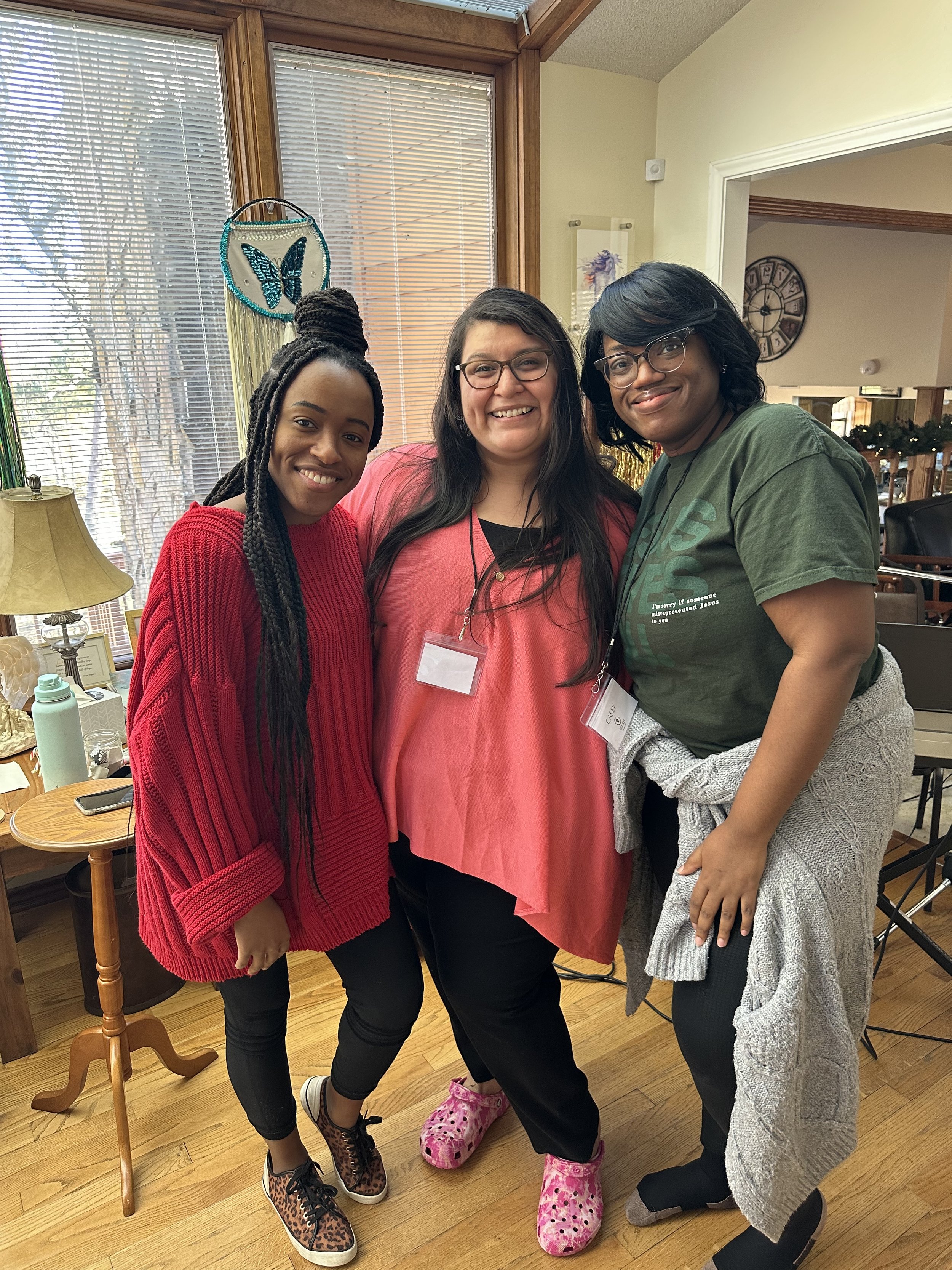 Three women smiling and posing together indoors. Two women are wearing glasses, and one has long twlined hair, while the woman in the middle has long dark hair. The woman on the left is wearing a red sweater and leopard-print shoes, the woman in the 