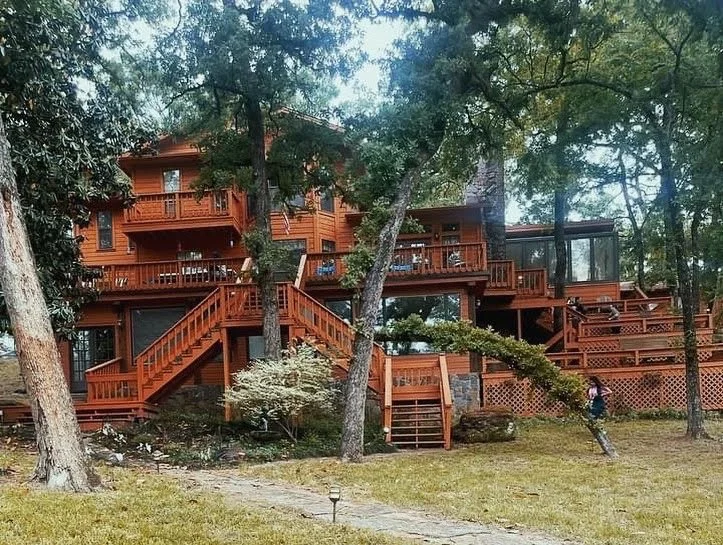 Large wooden house in a wooded area with multiple decks and stairs, surrounded by trees and a grassy yard.