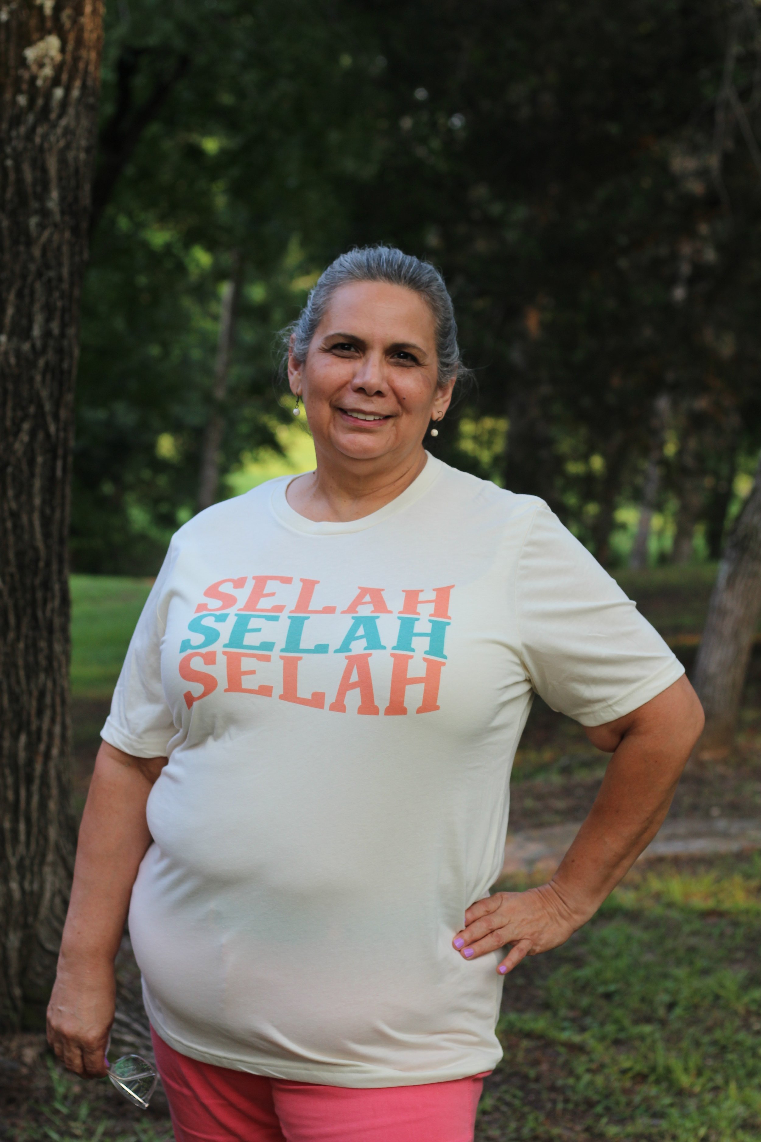 A woman standing outdoors near trees, wearing a cream T-shirt with 'Selah' written multiple times in orange, teal, and peach, smiling at the camera.