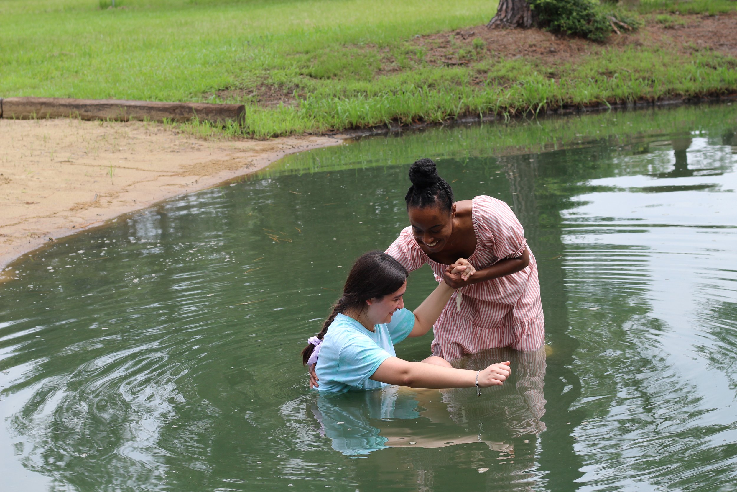 A woman helping another woman stand up in a pond or small lake, with trees and grass in the background.
