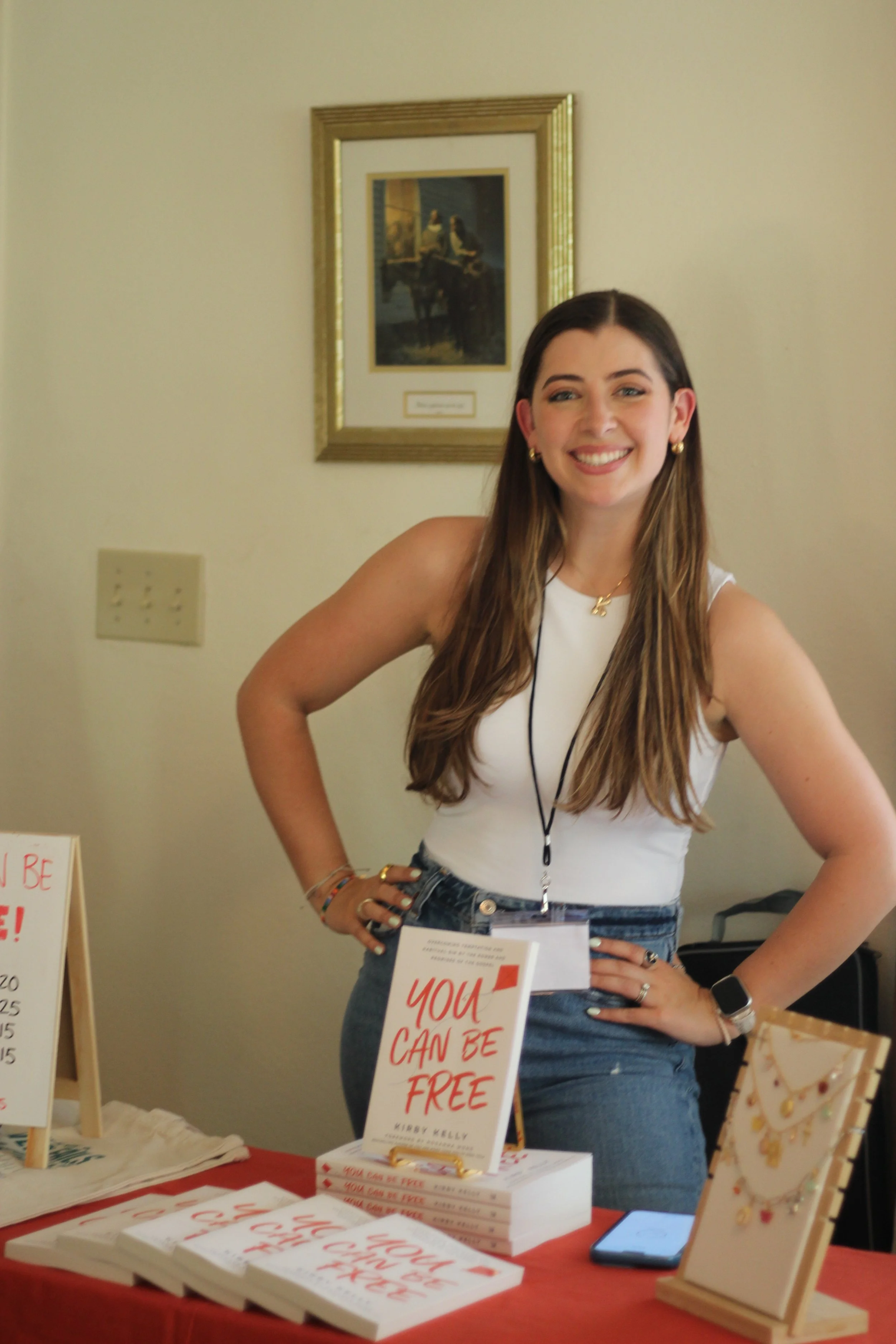 Young woman with long hair smiling at a table displaying books titled 'You Can Be Free' and jewelry, wearing a white tank top, jeans, a smartwatch, and accessories, standing indoors.