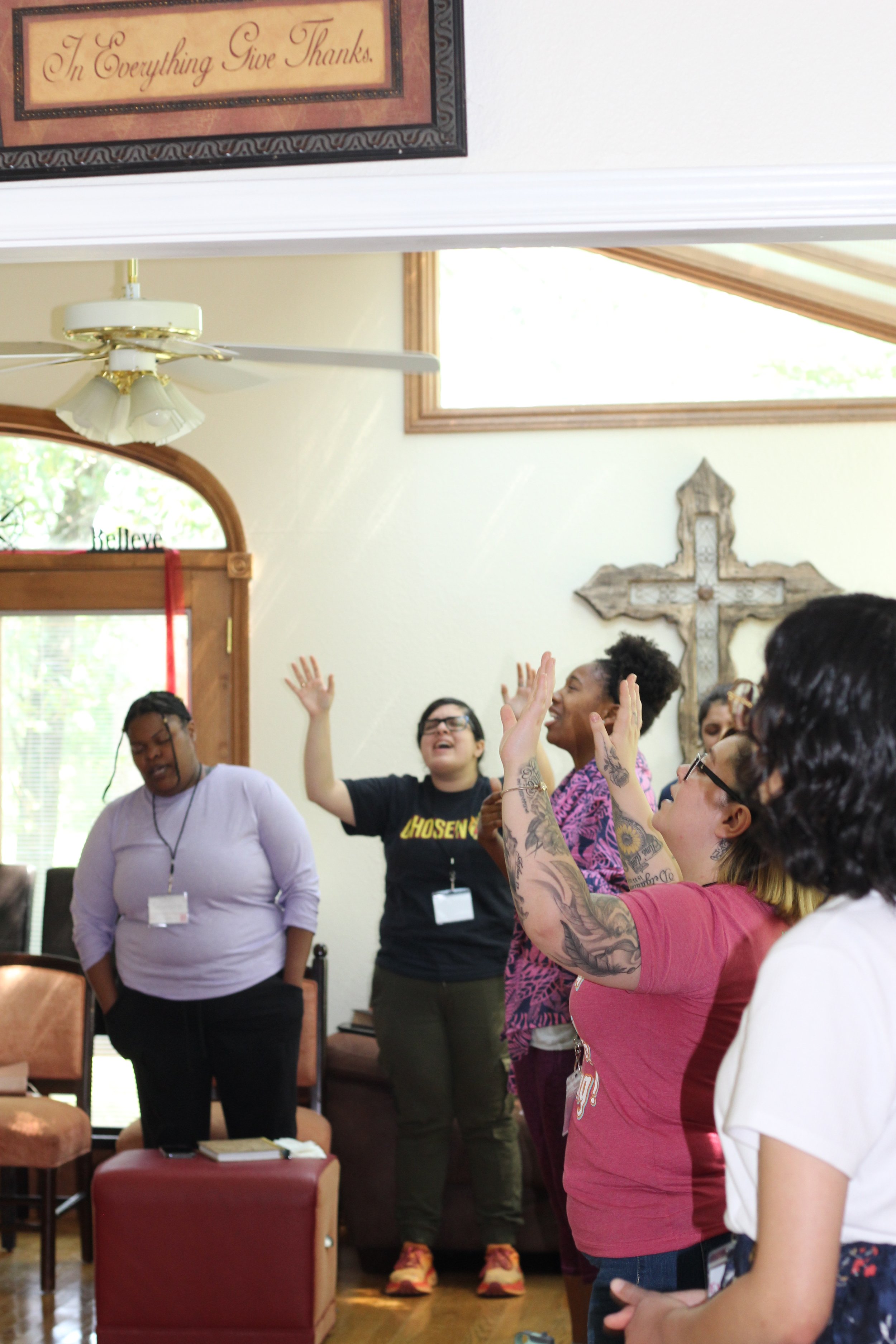 A group of women standing in a room with light-colored walls, some with hands raised, engaged in a joyful activity. One woman with tattoos and glasses has her hands raised, while others are smiling or with eyes closed, near a large wooden cross on th