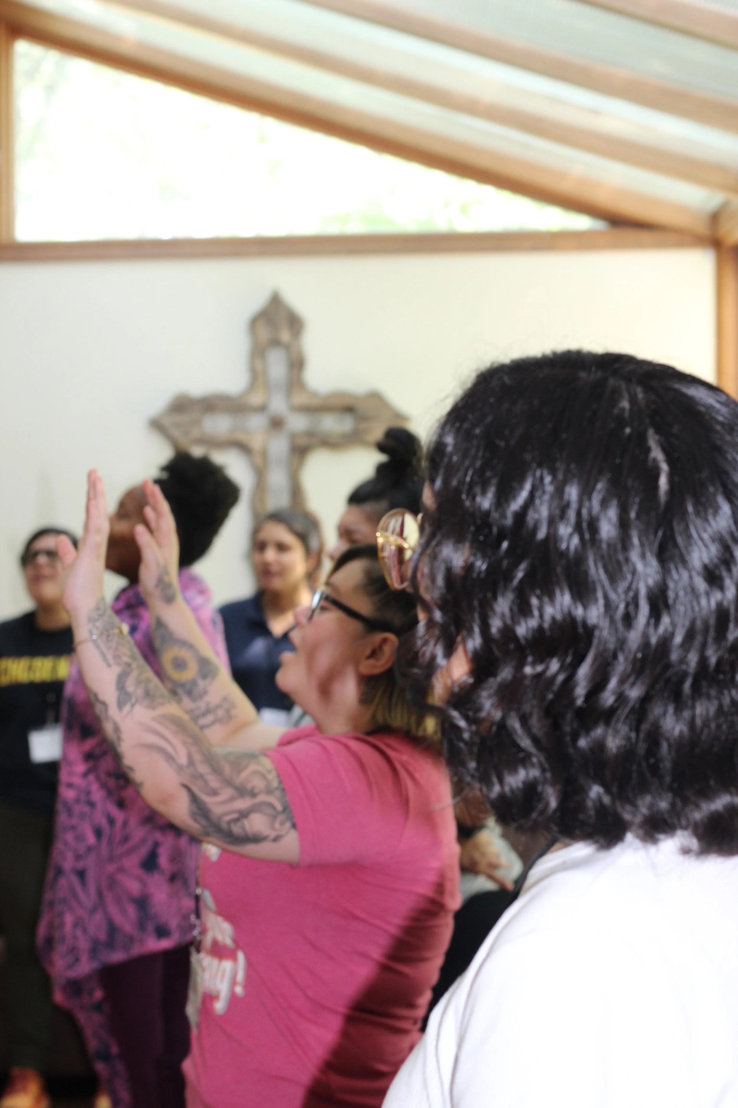 Group of people gathered indoors, some raising their hands, with a cross on the wall in the background.