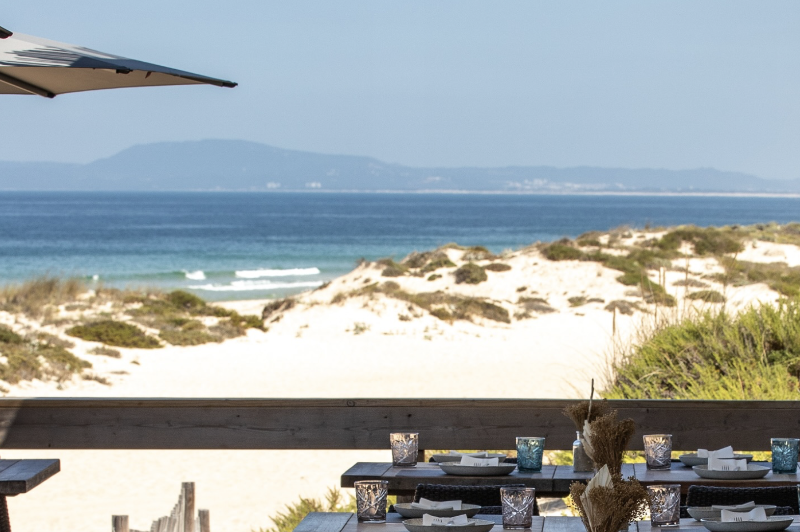 Empty outdoor dining table set with glasses and plates overlooking a sandy beach, dunes, and ocean under a clear sky.