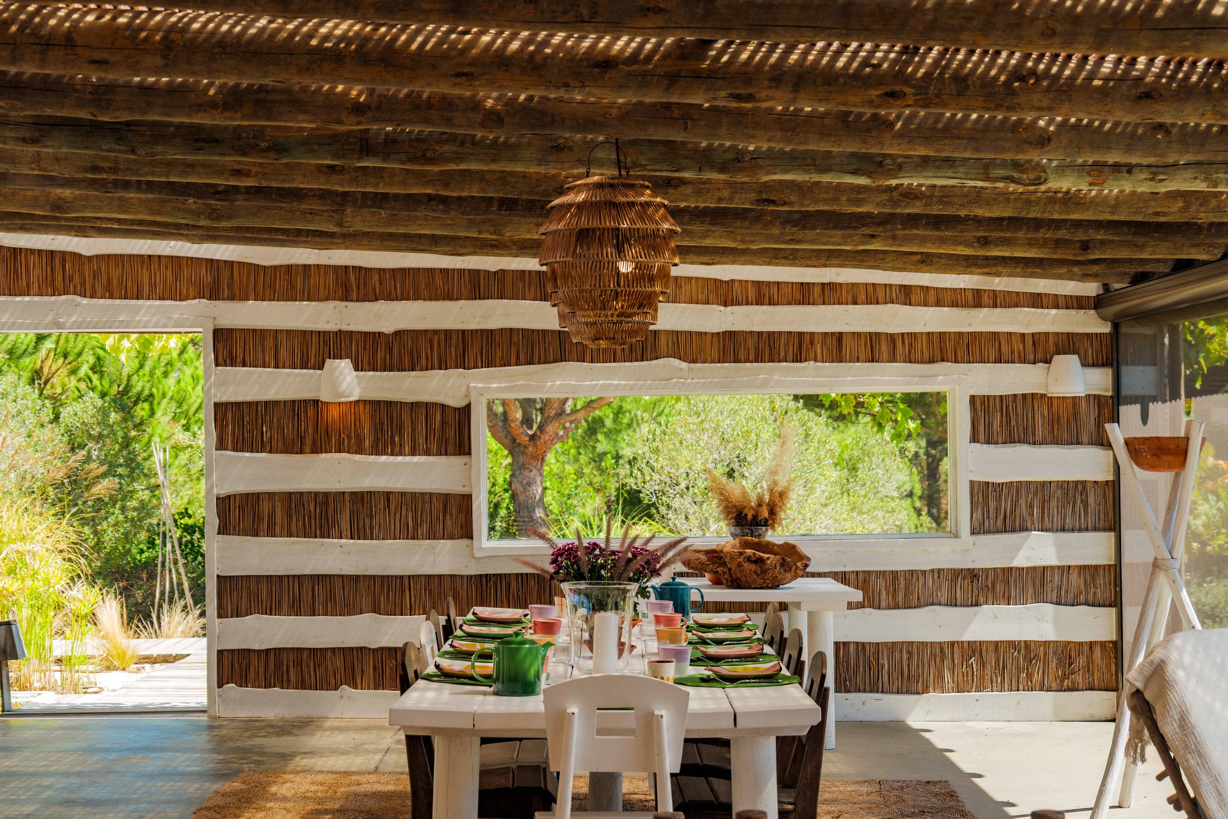A rustic dining room with a white wooden table set for a meal, with colorful mugs, plates, and a floral centerpiece. The room has a natural, earthy decor with a thatched ceiling, wooden beams, and walls made of horizontal wooden slats. Large windows 