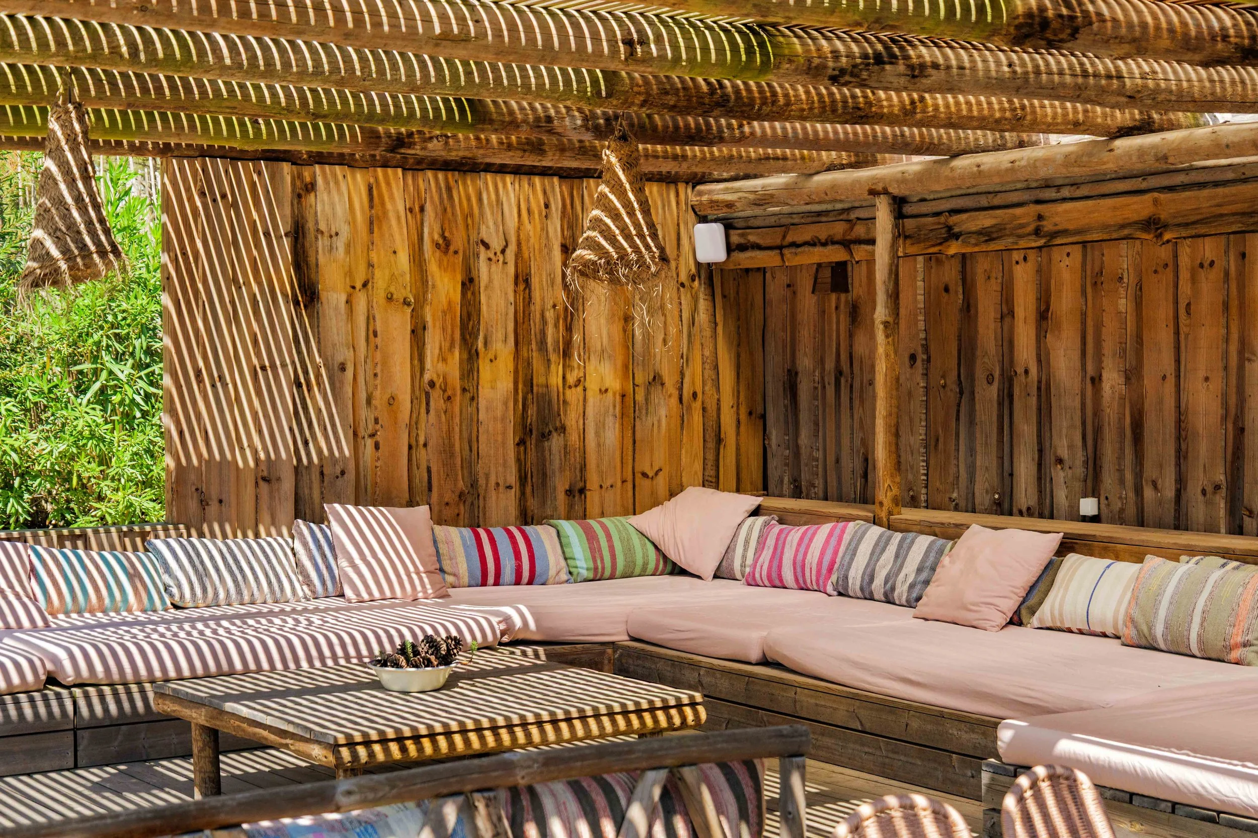 Outdoor seating area with a corner sofa with pink cushions, colorful striped pillows, and a wooden table, shaded by slatted wood and rope decorations, surrounded by green foliage.