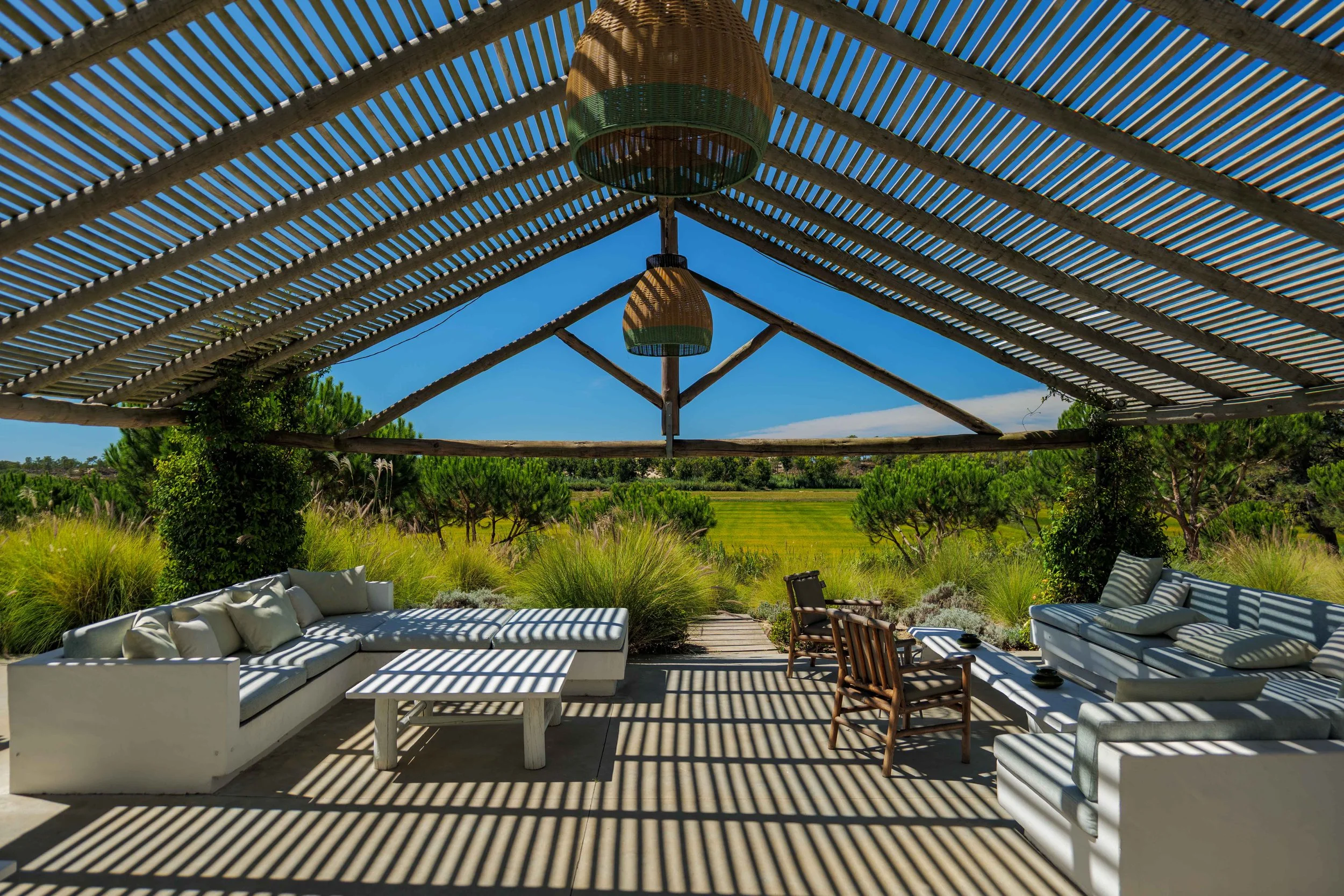 Outdoor patio with white furniture, wooden chairs, and striped cushions under a pergola with bamboo slats, overlooking green fields and trees on a sunny day.