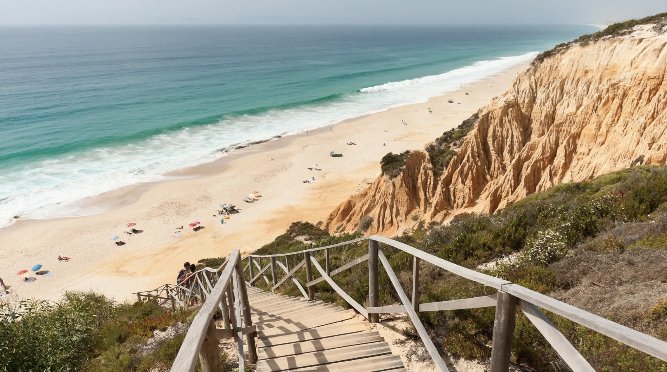 Wooden staircase leading down to a sandy beach with umbrellas, people, and ocean waves, with rocky cliffs to the side and greenery at the top.