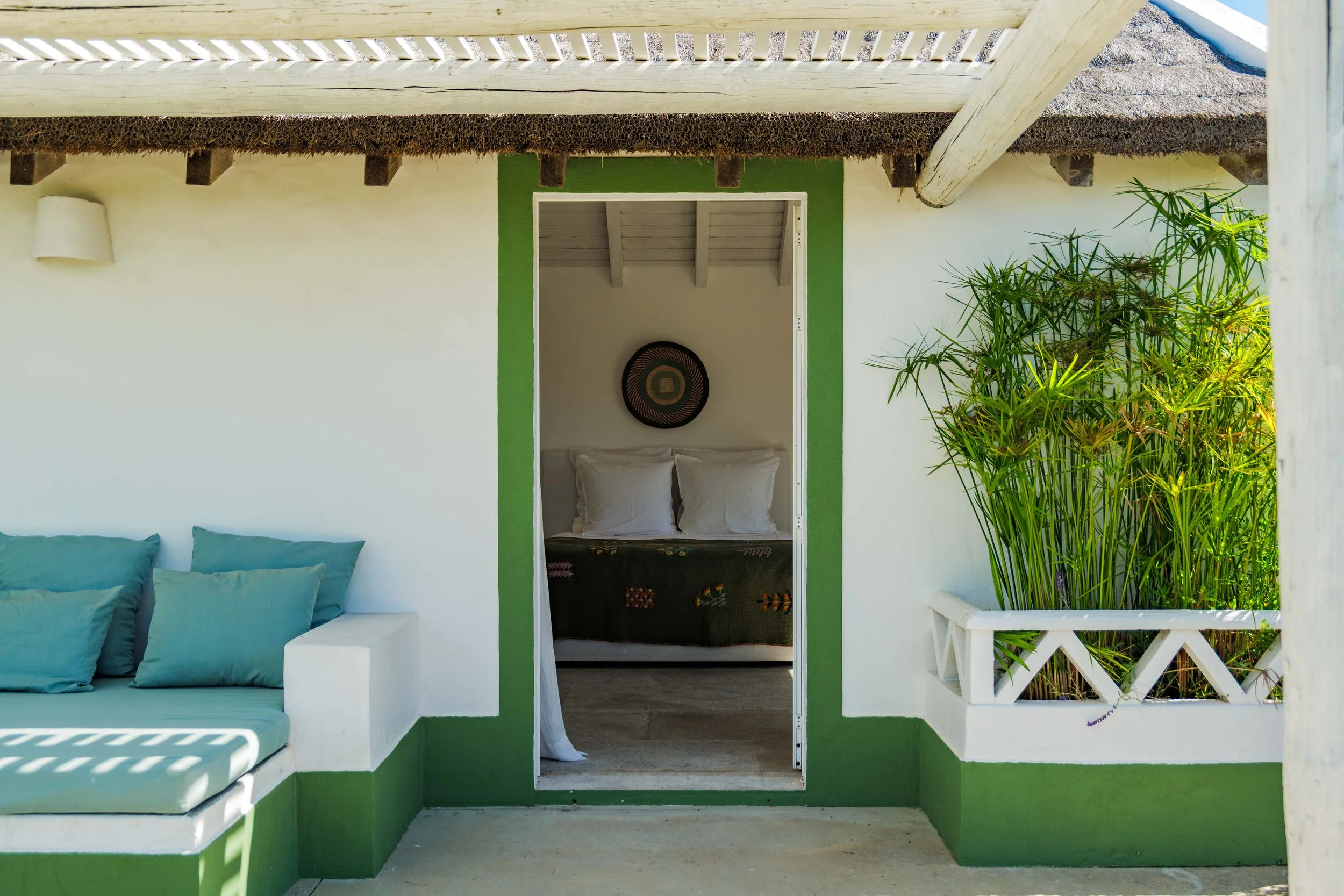 View of a small bungalow with white walls, green trim around the door, and a thatched roof. The front porch has a built-in seating area with teal cushions on the left, and a doorway leading into a bedroom with white pillows and dark bedspread inside.