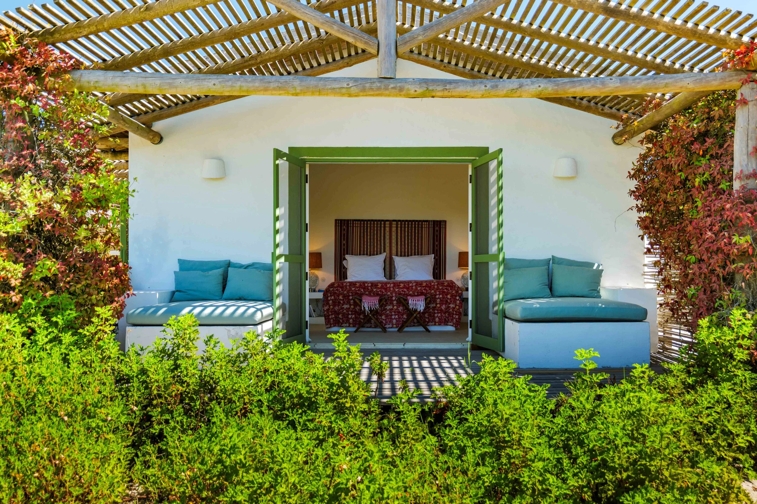 A cozy outdoor patio with white walls, green shutters, and a wooden lattice roof. The patio has built-in white seating with blue cushions and is surrounded by lush green bushes and red flowering plants.