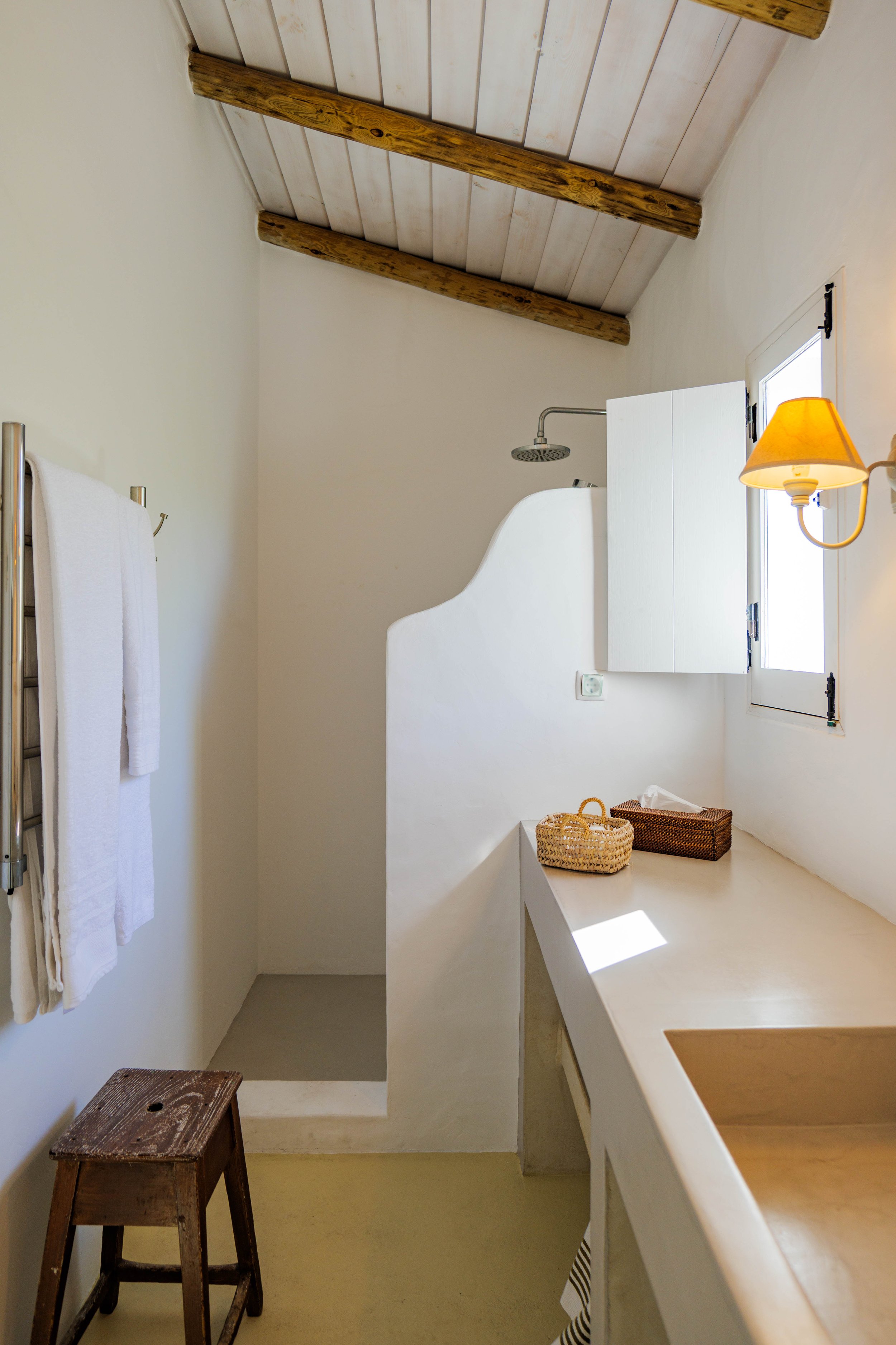 A cozy bathroom with white walls, a wooden ceiling with exposed beams, a small window, a wall-mounted lamp, a shower area with a showerhead, a towel rack with white towels, a wooden stool, a countertop with a wicker basket and tissue box, and a partial view of a sink.