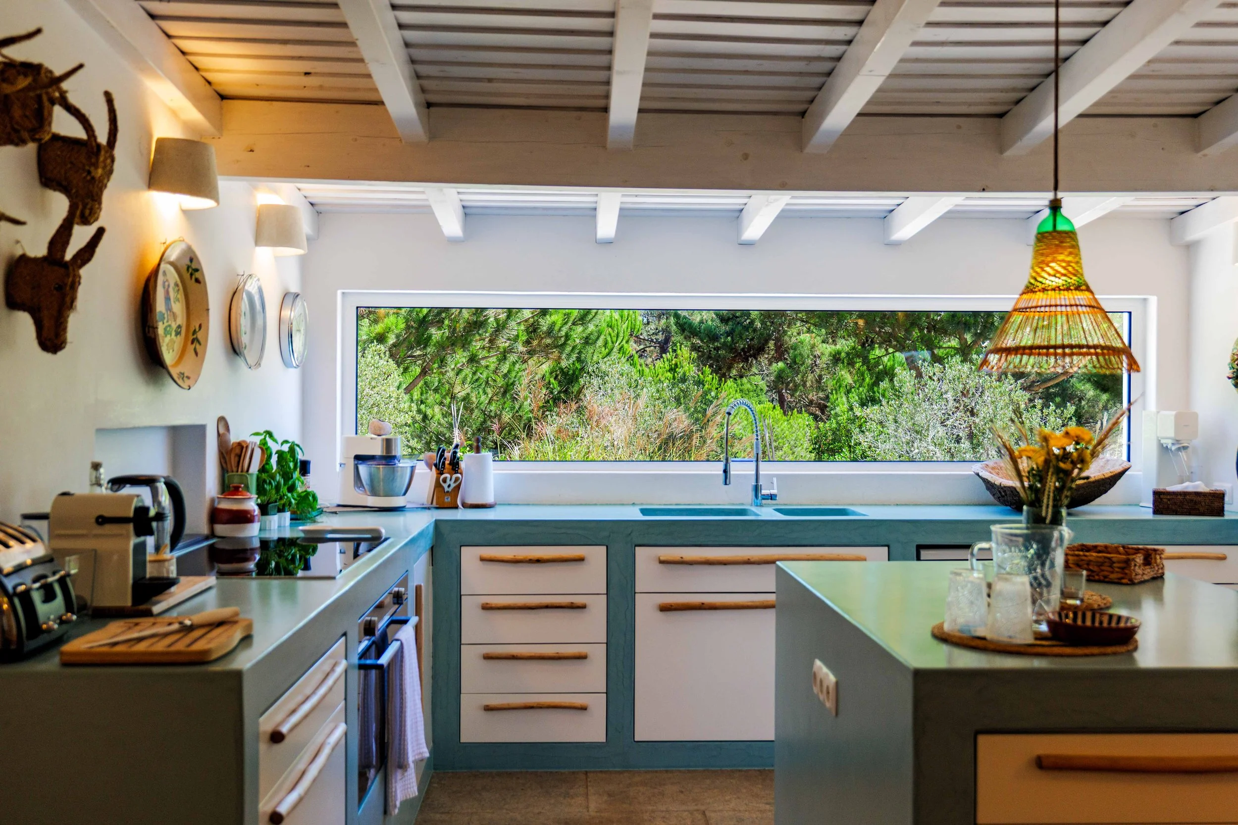 Modern kitchen with large window overlooking green trees, featuring white cabinets with wooden handles, kitchen appliances, and decorative wall plates.