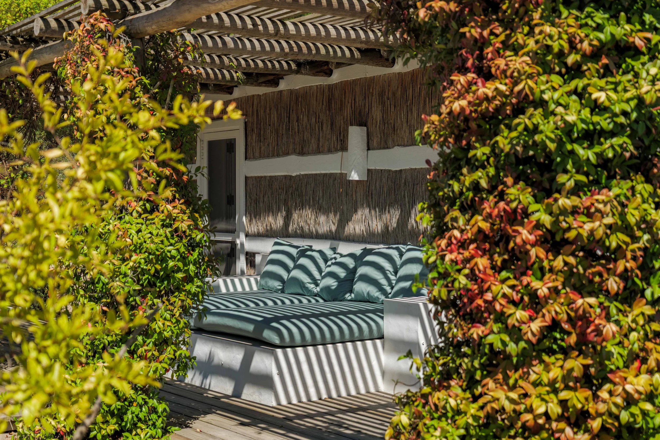 A cozy outdoor seating area with a white couch and light blue cushions, surrounded by lush green and red foliage, with sunlight casting striped shadows on the furniture.