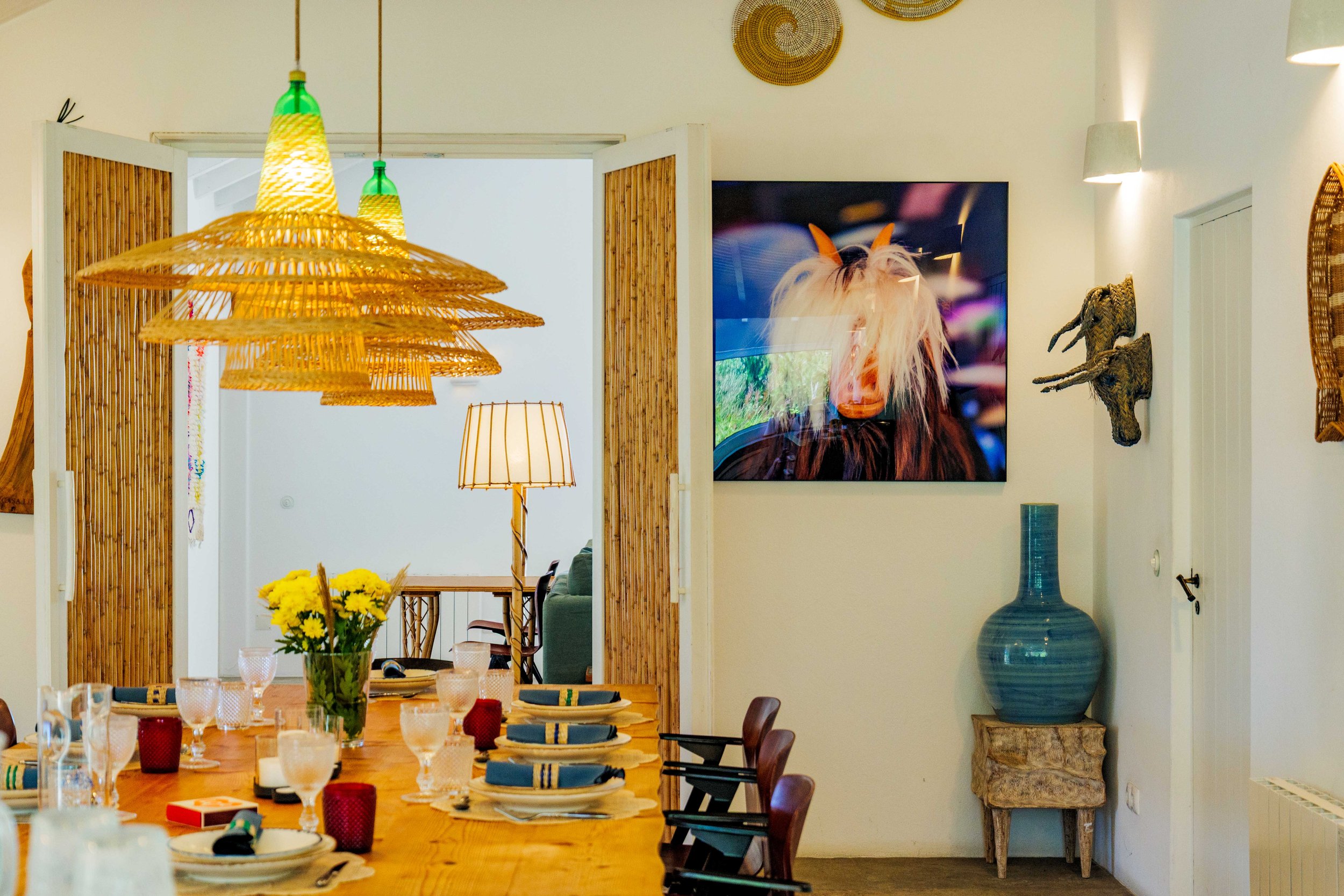 A dining room with a wooden table set for a meal, with a centerpiece of yellow flowers. Overhead several large wicker pendant lights. The background features a part of a living room with a green couch, a table, and a lamp. The walls are decorated wit