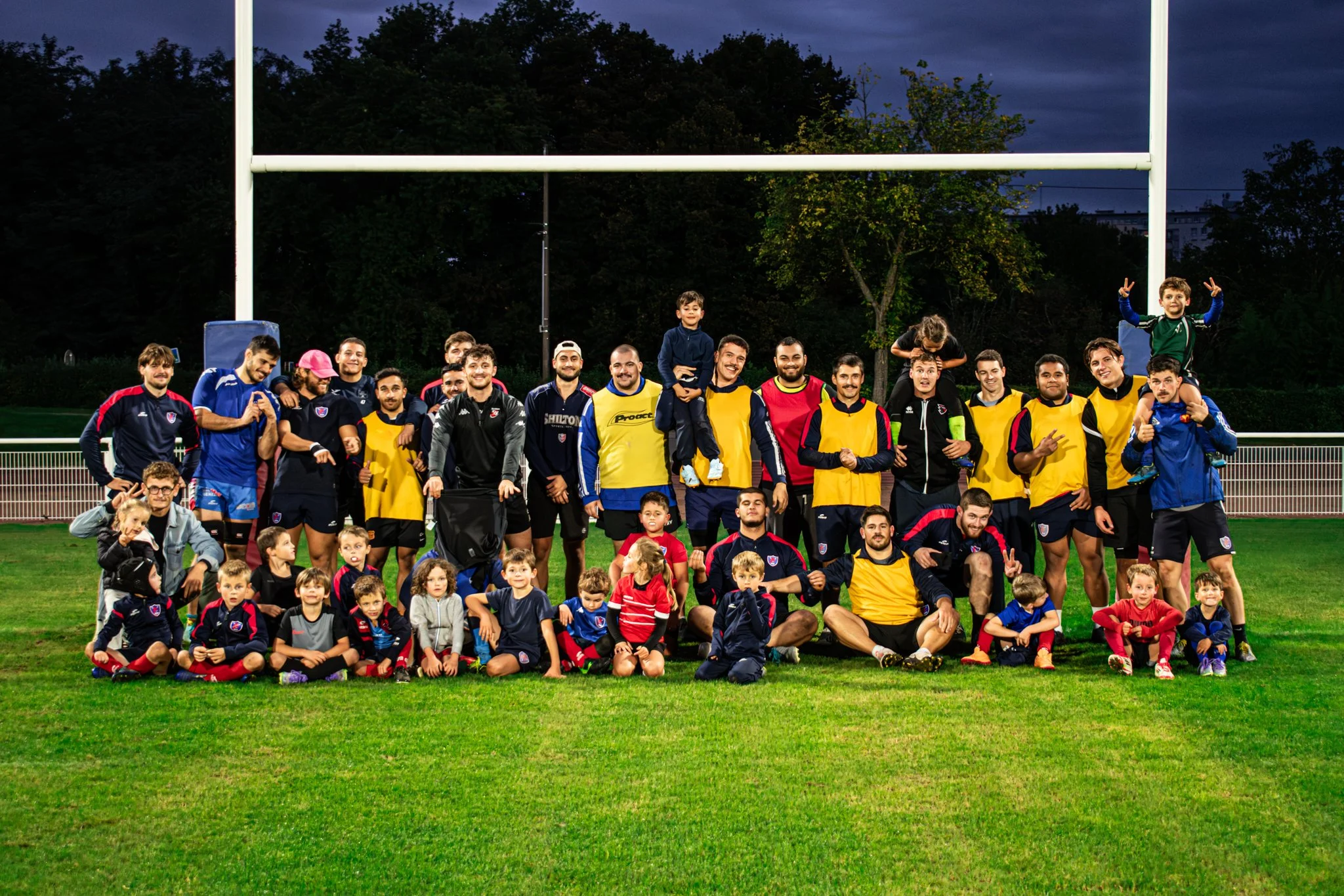 Une grande famille de joueurs de rugby, hommes, femmes et enfants, posant sur un terrain en plein air lors d'une séance photo en soirée, avec un but de rugby derrière eux.