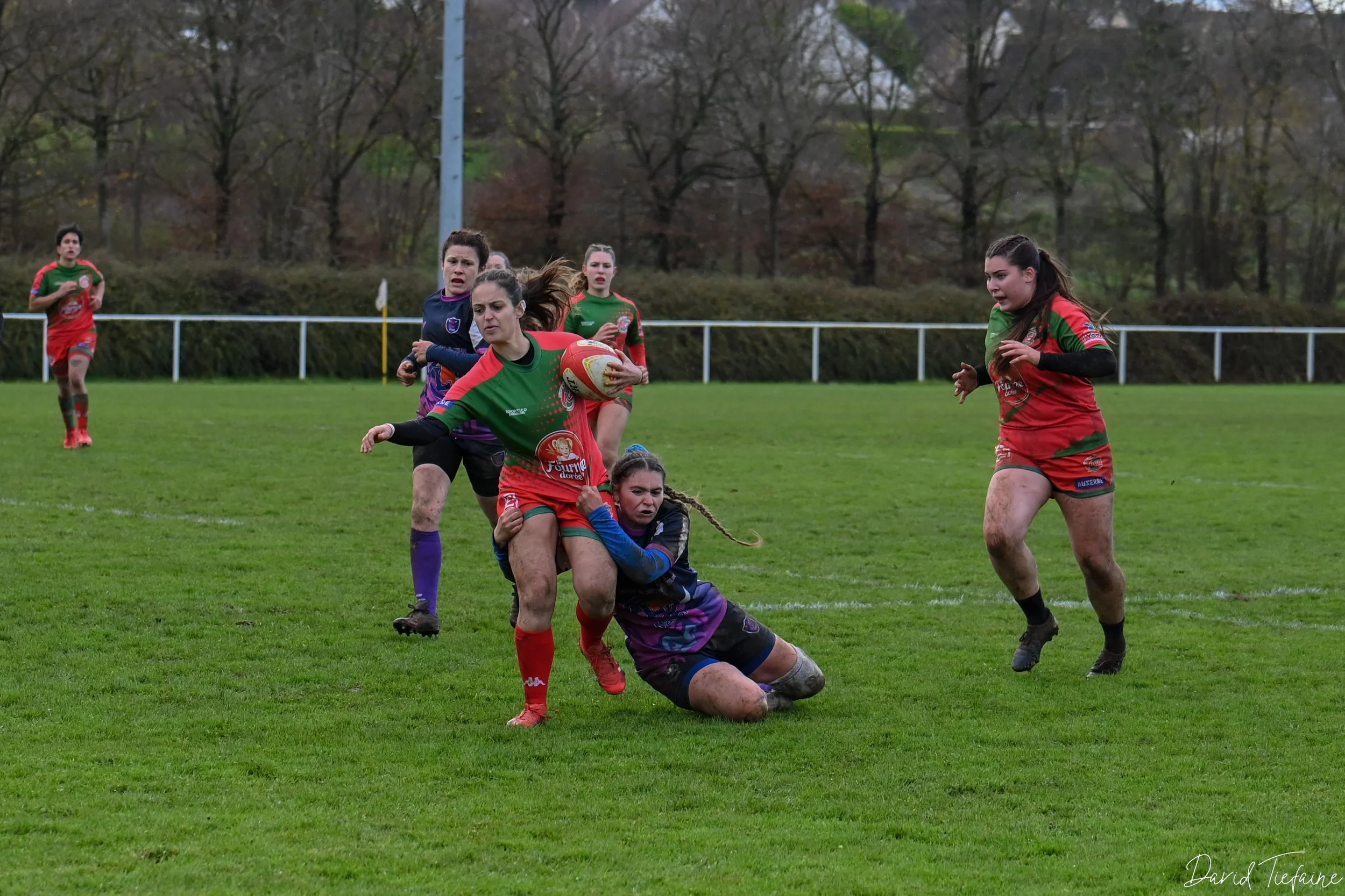 Femme jouant au rugby, tenant le ballon, en train d'être plaquée par une autre joueuse, dans un terrain de rugby avec d'autres joueuses en arrière-plan.