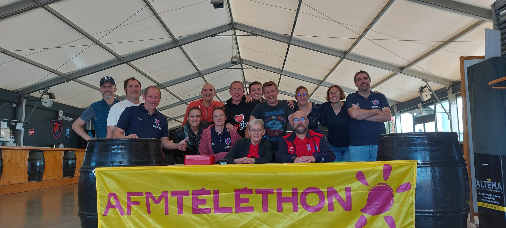 Groupe de personnes réunies sous une tente, posant pour une photo lors d'un événement de collecte de fonds, avec une bannière jaune portant l'inscription 'AFM TELETHON' et deux grands tonneaux en plastique.