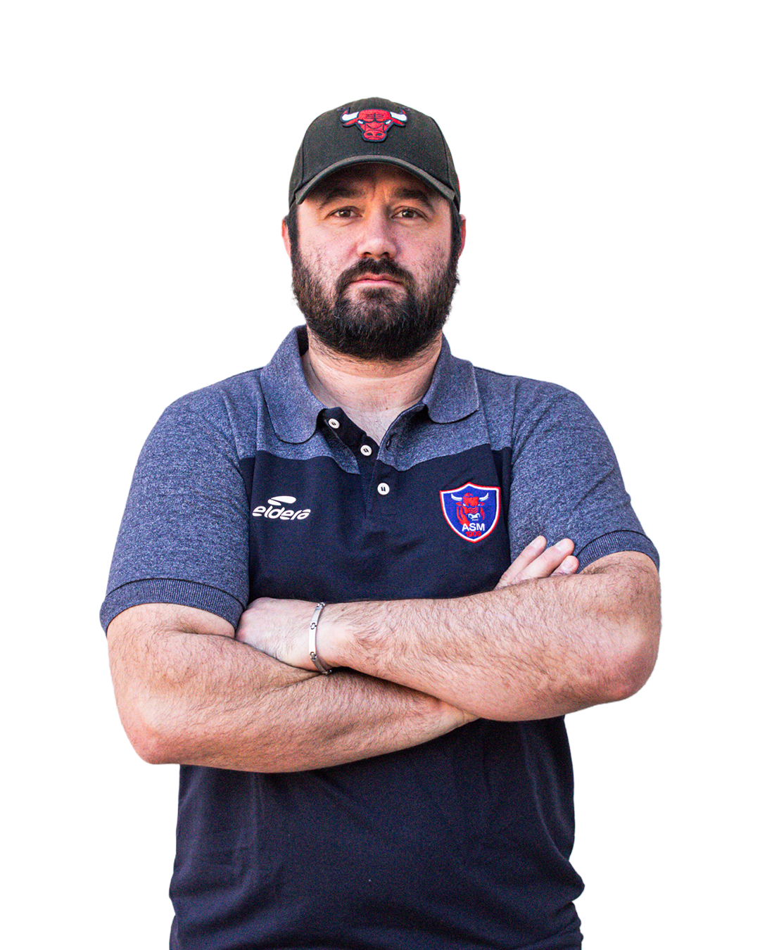 Un homme avec une barbe porte un polo gris avec du bleu marine et un logo d'équipe de football, et un bonnet noir avec un logo de taureau.