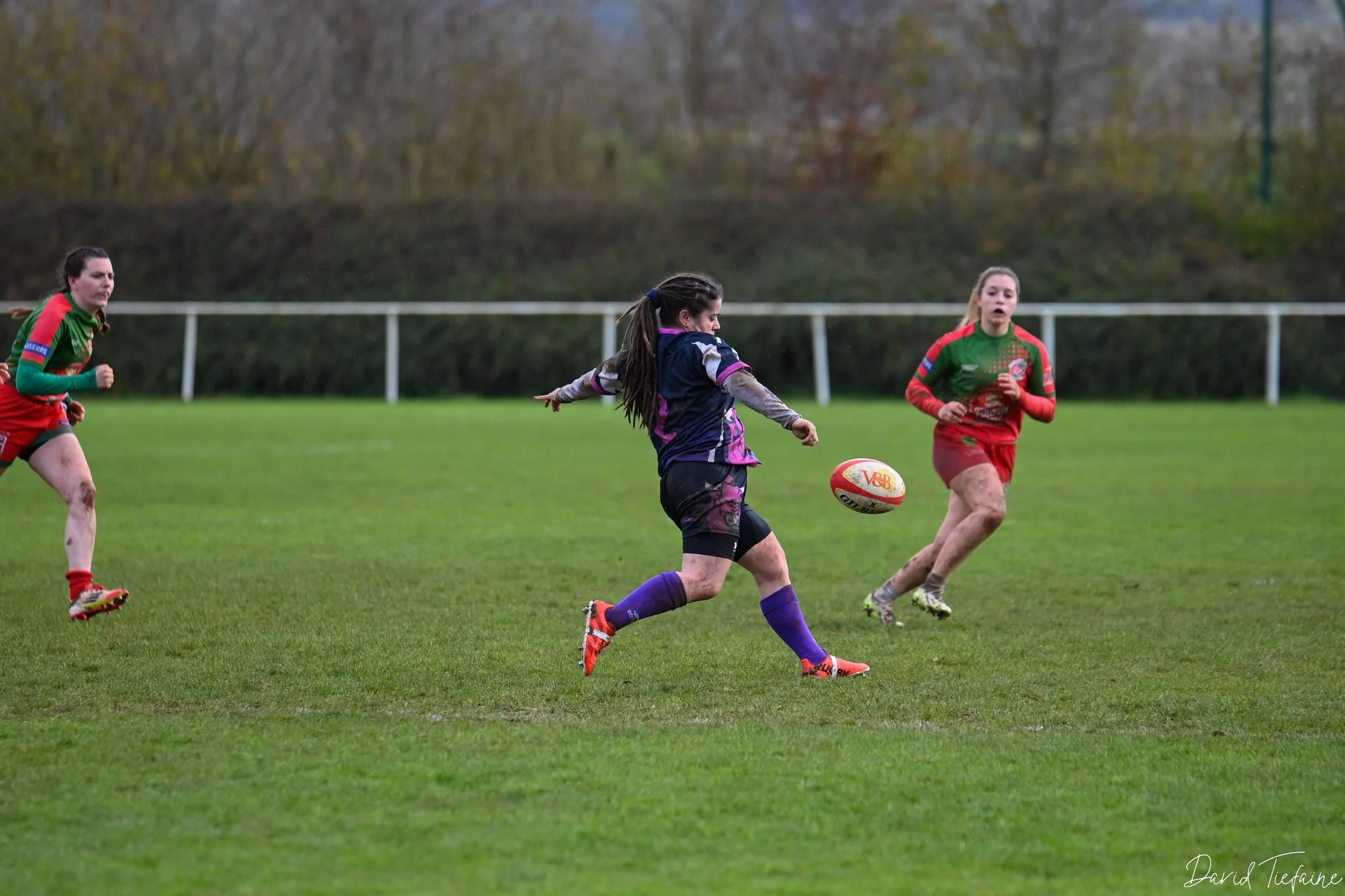 Trois femmes jouant au rugby sur un terrain vert, deux en maillots verts et rouges et une en maillot noir et violet, en pleine action pour attraper ou envoier le ballon.