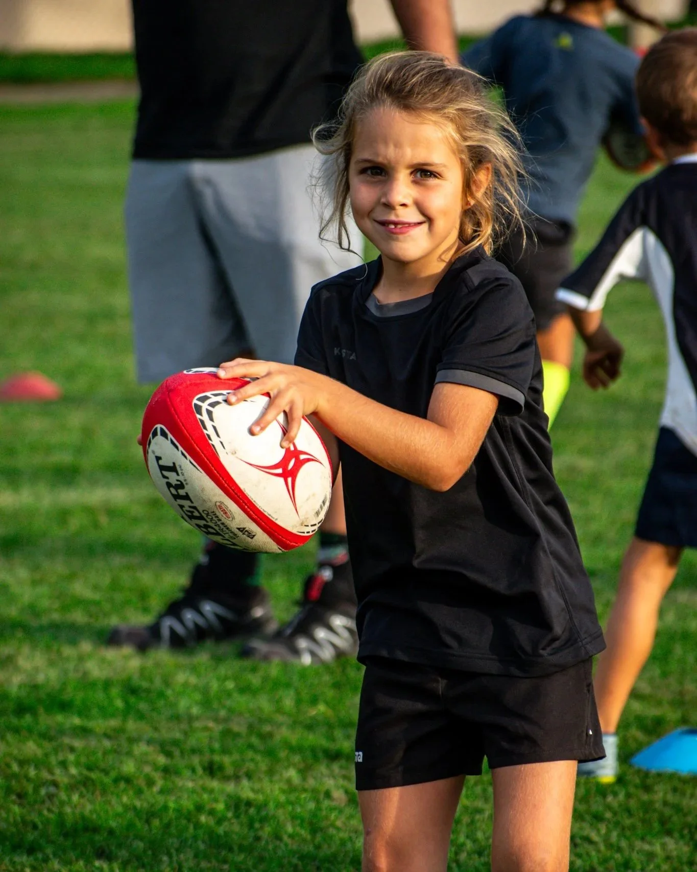 Une jeune fille tient un ballon de rugby sur un terrain de sport, portant une tenue de sport noire, avec d'autres enfants et adultes en arrière-plan.