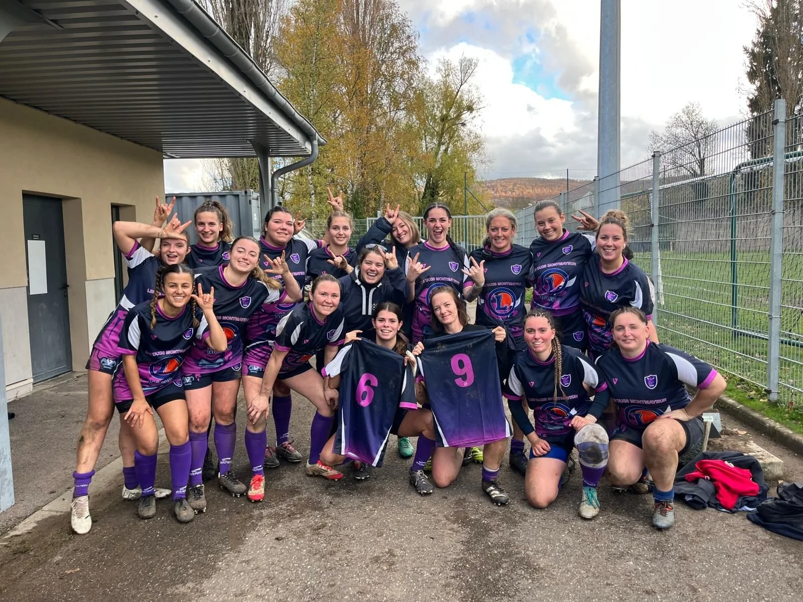 équipe féminine de rugby posant ensemble après un match, certains tenant des maillots avec les numéros 6 et 9, sous un ciel nuageux avec des arbres d'automne en arrière-plan