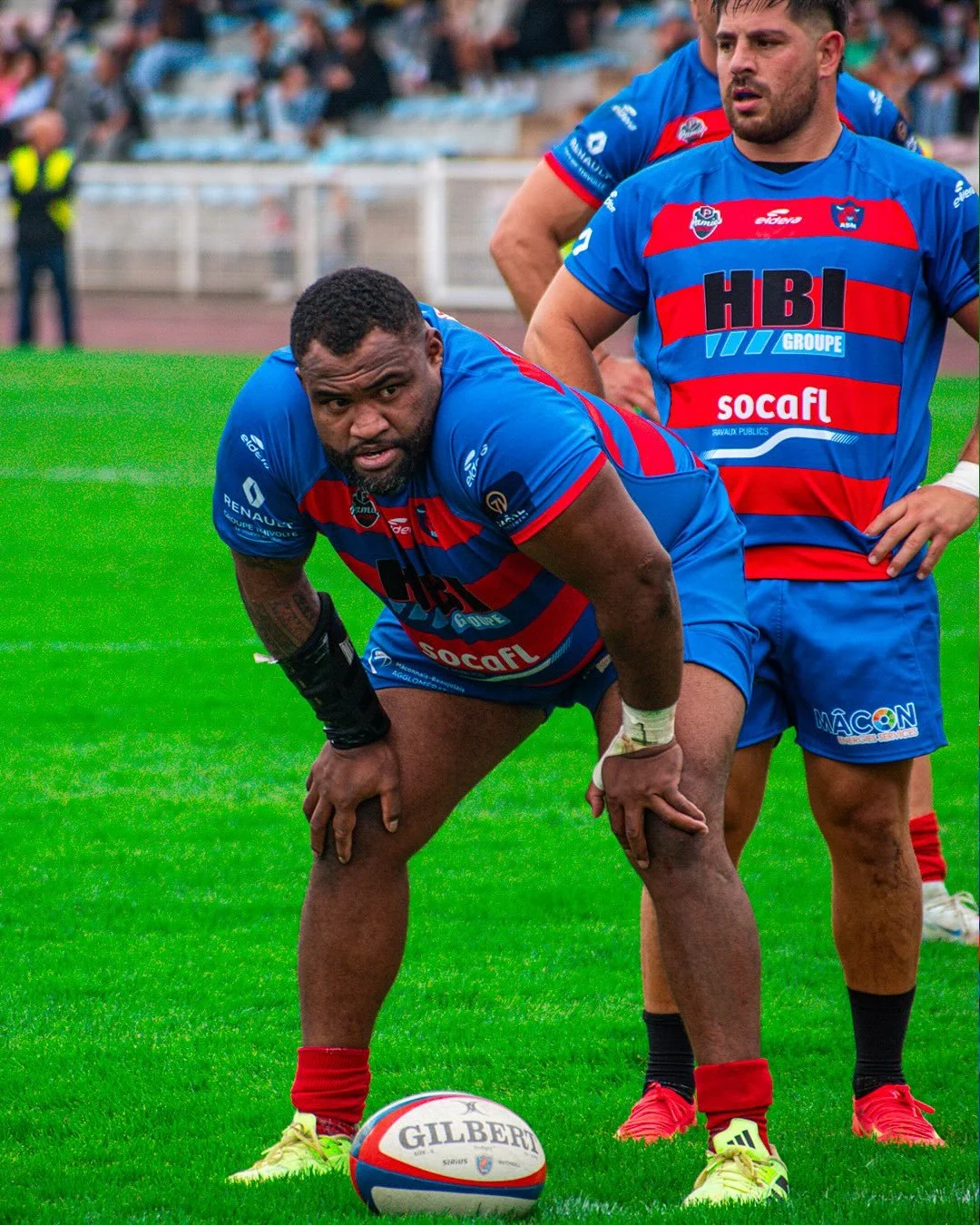 Deux joueurs de rugby en uniforme bleu avec rayures rouges, sur un terrain vert lors d'un match, dont un est penché sur un ballon.