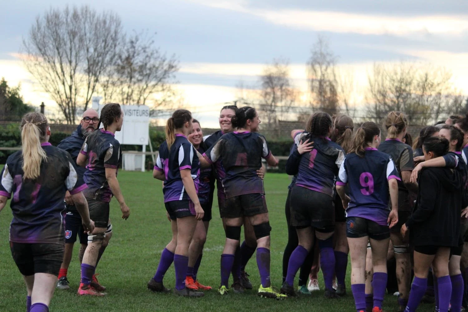 Une équipe de rugby féminine en tenue violette et noire, souriante et en groupe sur un terrain en fin de match ou entraînement, avec un ciel nuageux en arrière-plan.