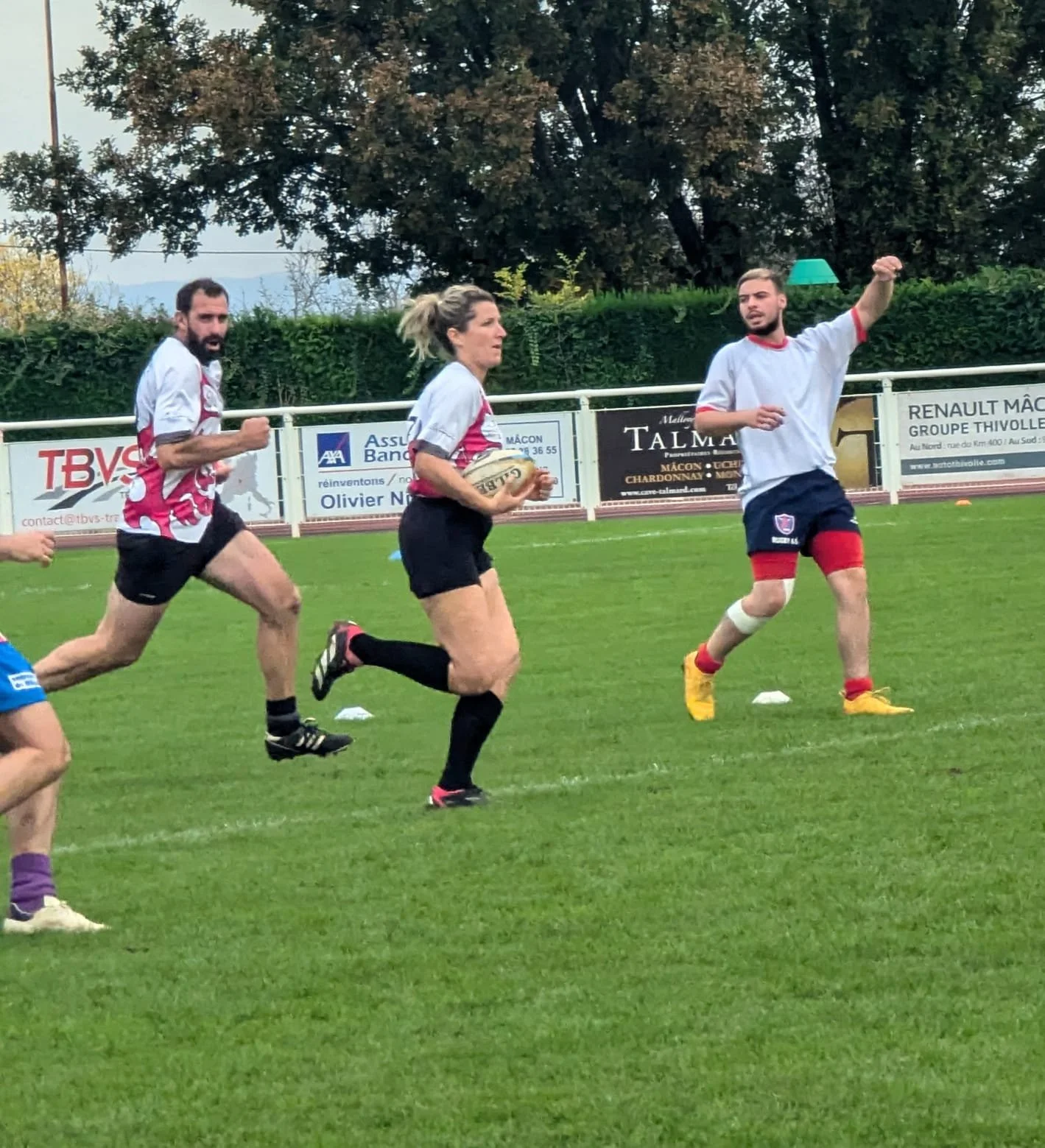 Des joueurs de rugby en action sur un terrain vert, trois portant des maillots blancs et un avec un maillot bleu, sous un ciel largement dégagé.