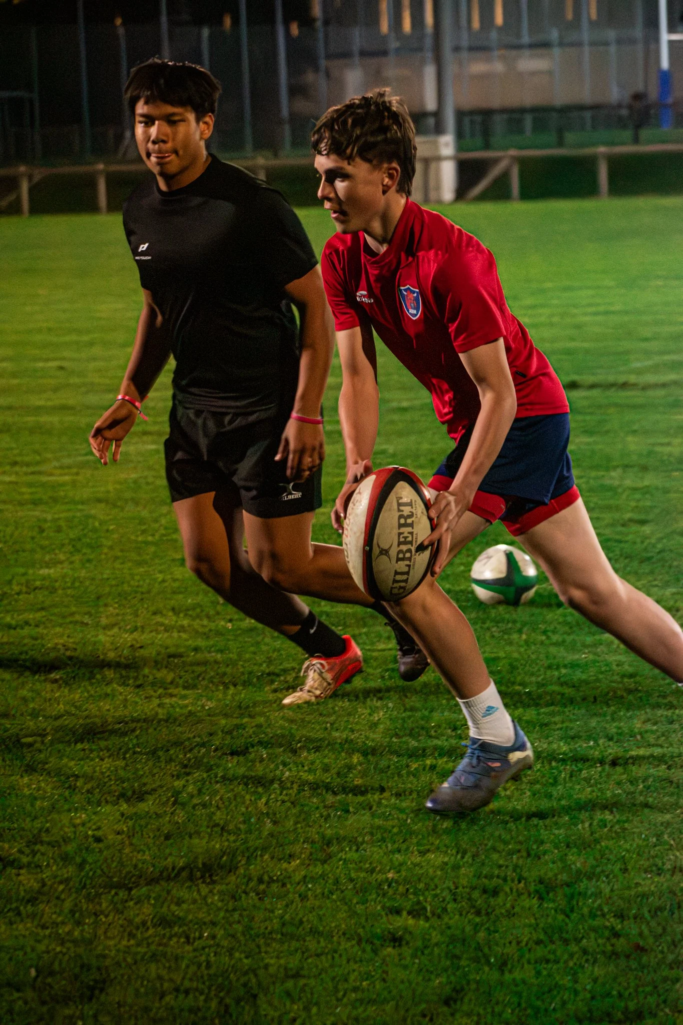 Deux jeunes rugbymen jouant sur un terrain en gazon, un en maillot noir et l'autre en maillot rouge, avec un ballon de rugby en main, lors d'une séance d'entraînement ou d'un match de rugby.