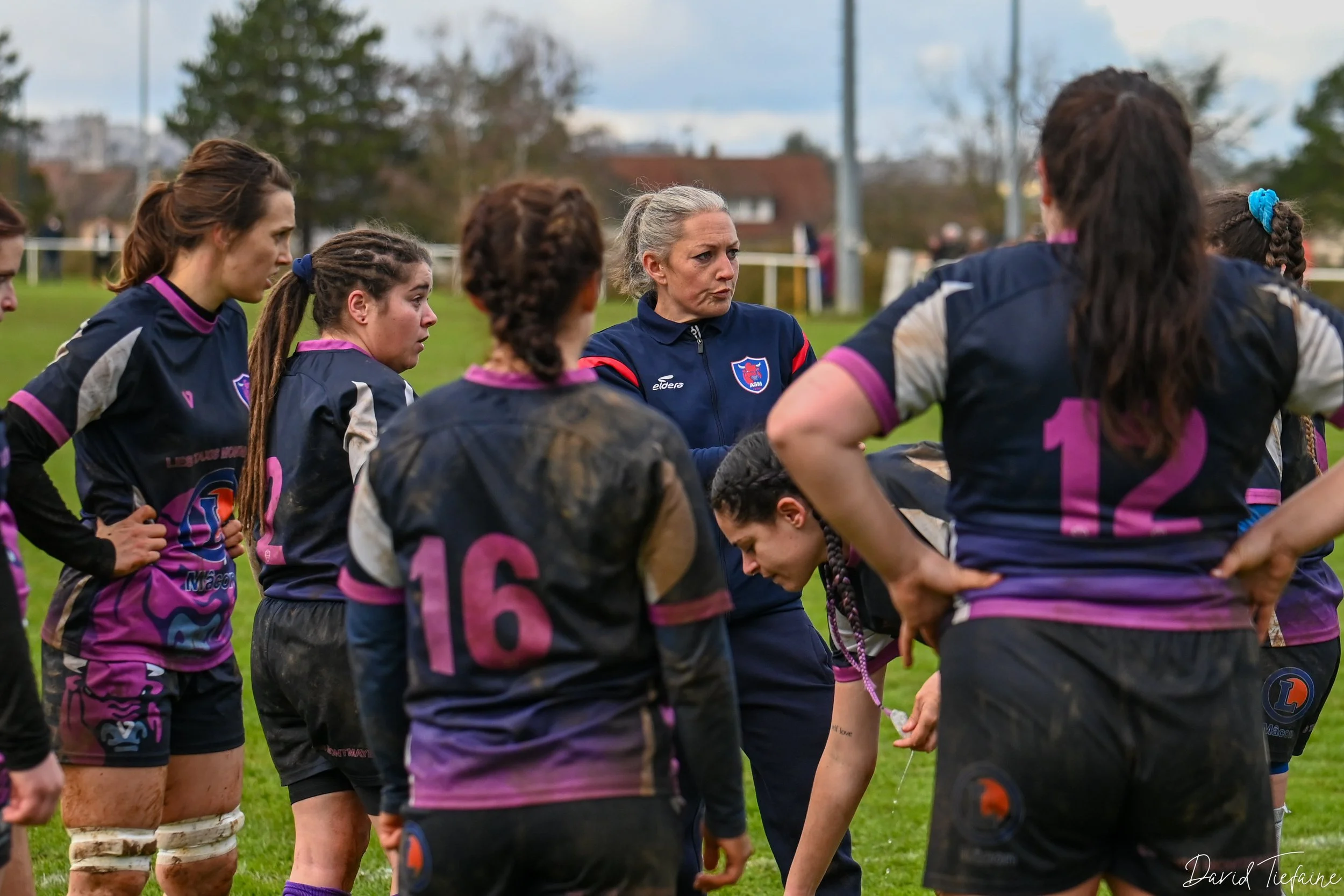 Une équipe féminine de rugby écoute leur entraîneure pendant une séance d'entraînement sur un terrain herbeux, avec des vêtements de sport nâtés et une ambiance concentrée.