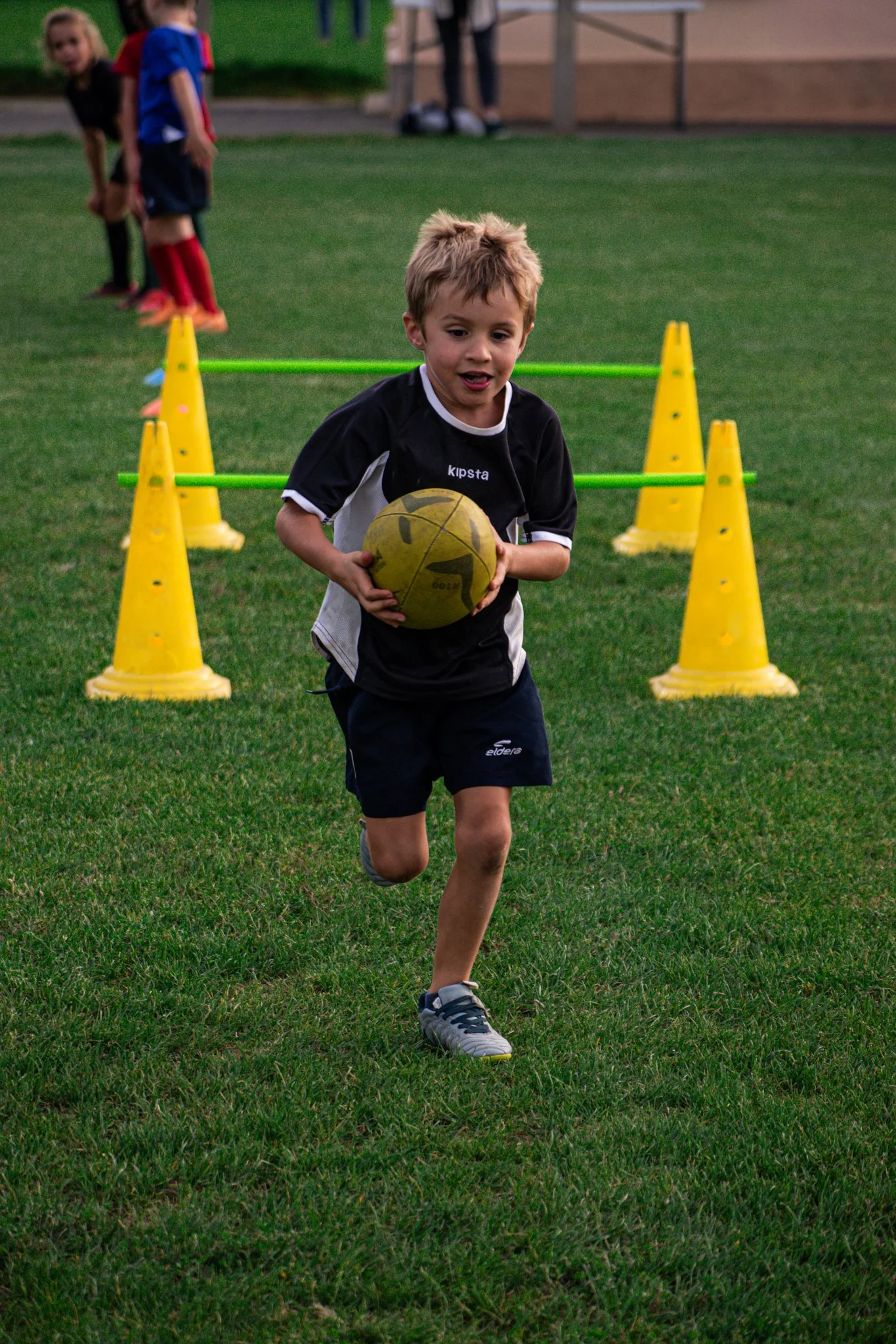 Un jeune garçon court sur un terrain d'herbe en tenant un ballon de rugby, avec des cônes jaunes et des barres pour un entrainement sportif, sur un fond de groupes d'enfants.