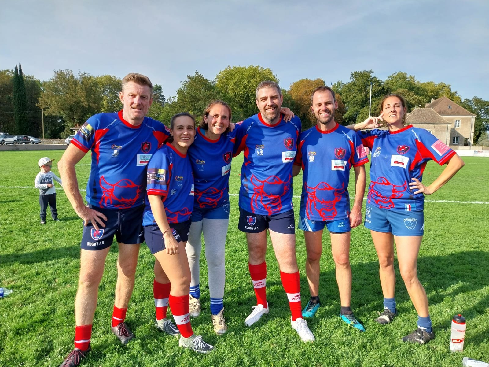 Groupe de sept personnes portant des maillots de rugby bleus avec un lion rouge sur le devant, posant sur un terrain herbeux, avec des arbres et une maison en arrière-plan.
