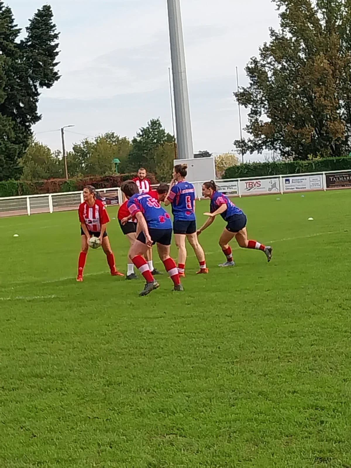 Joueuses de rugby en action sur un terrain en plein air, avec deux équipes opposées, une en rouge et blanche, l'autre en bleu et rouge, lors d'un match ou entraînement.