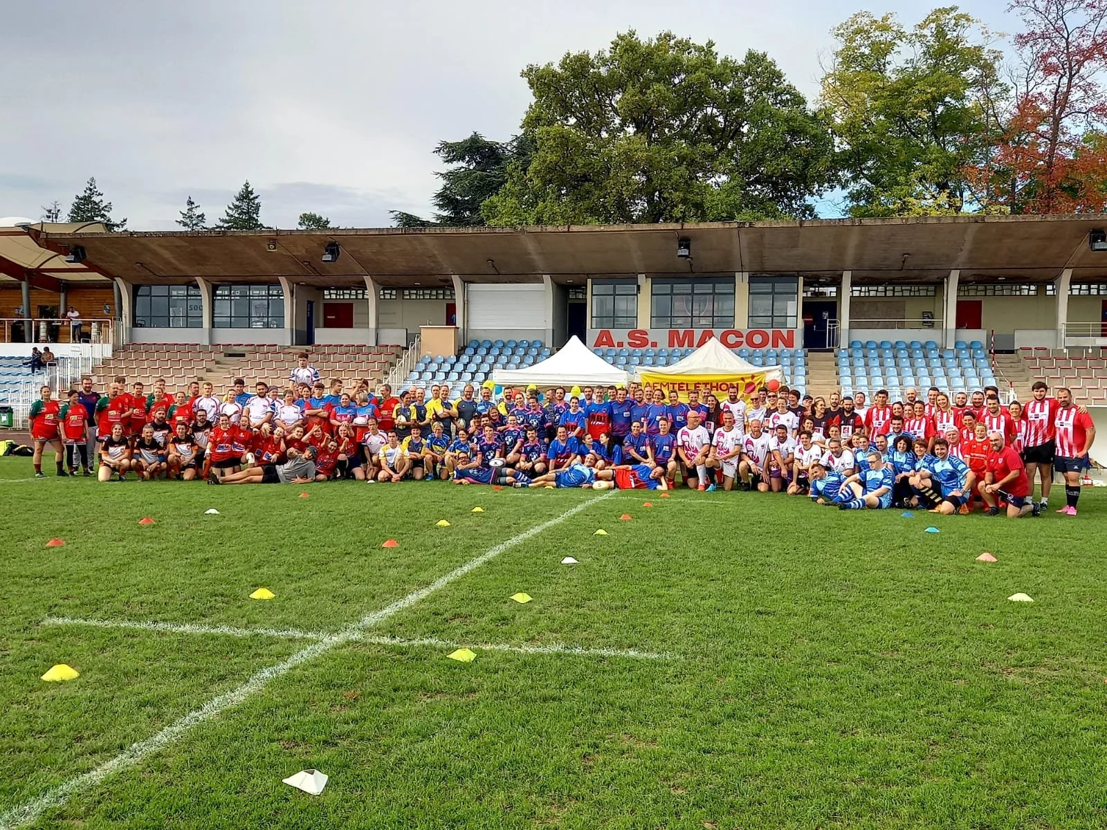 Un grand groupe de personnes debout et assises sur un terrain de sport, probablement après un match ou un événement sportif, avec des gradins en arrière-plan et des arbres. La majorité porte des maillots de rugby rouges, bleus ou blancs.