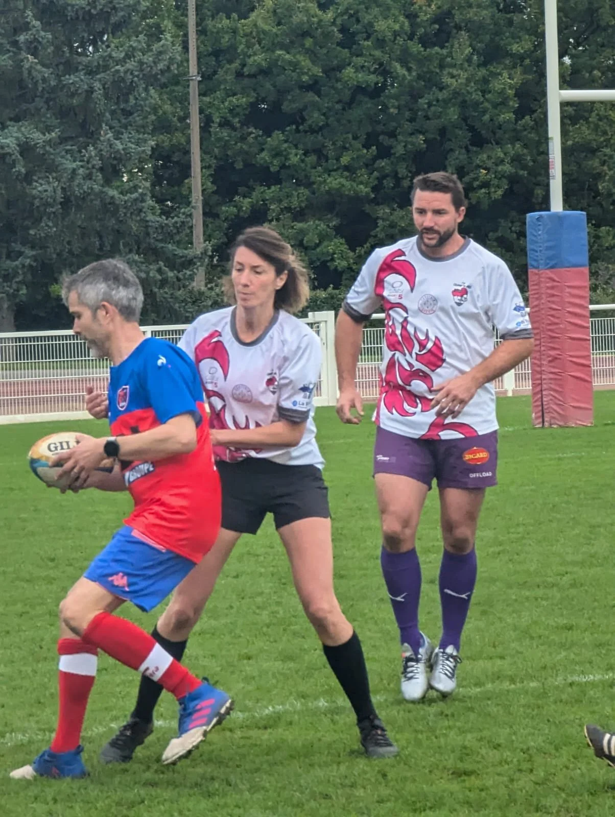 Trois personnes jouent au rugby sur un terrain en extérieur, deux hommes et une femme, avec un ballon ovale en main. Le fond montre beaucoup d'arbres.
