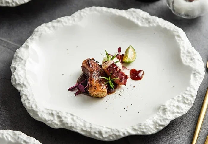 Elegant white textured ceramic plate with a small portion of seared meat, garnished with herbs, a lime wedge, and sauce drops.