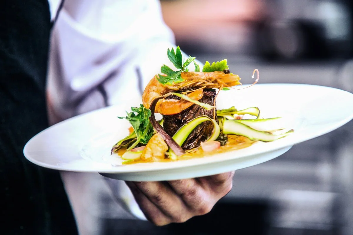 A chef holding a white plate with a gourmet seafood dish: a large shrimp, slices of cucumber, greens, and a dark sauce or broth.